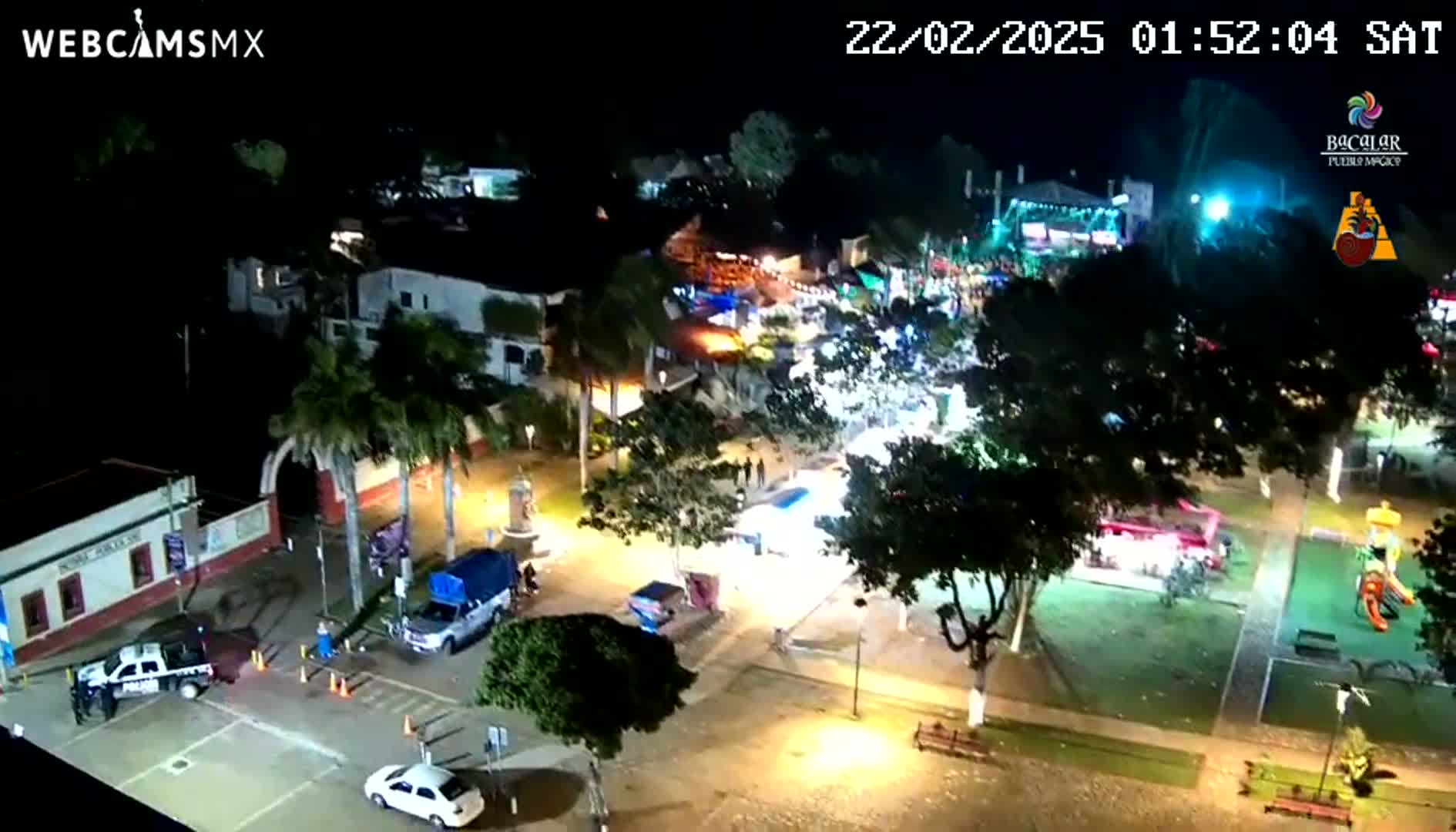 An aerial nighttime view of a town square with brightly lit buildings, vehicles, and people gathered in a plaza under a clear sky.