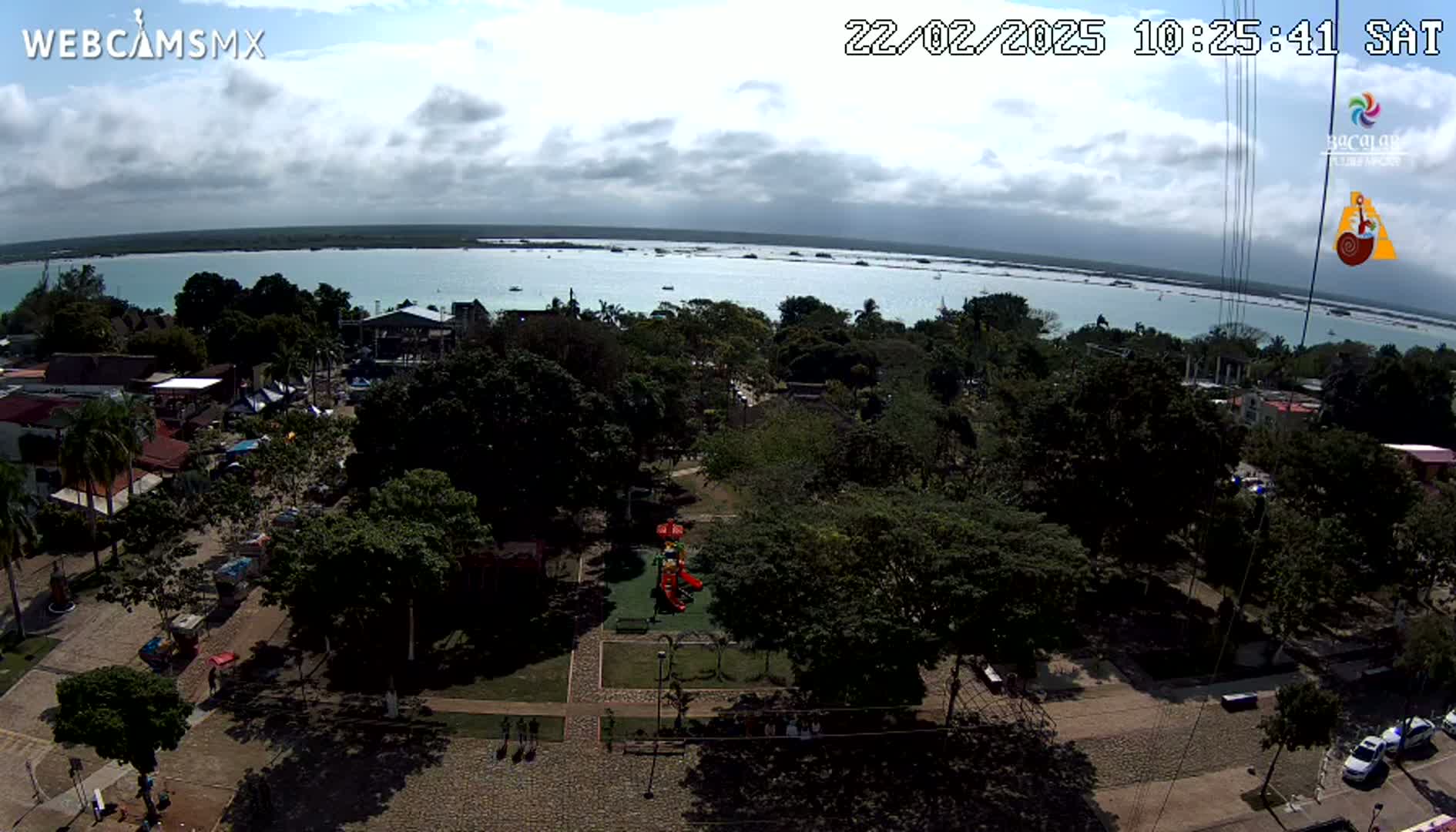 An aerial view shows a town square with a playground, surrounded by lush green trees, overlooking a calm, light-blue body of water under a partly cloudy sky.