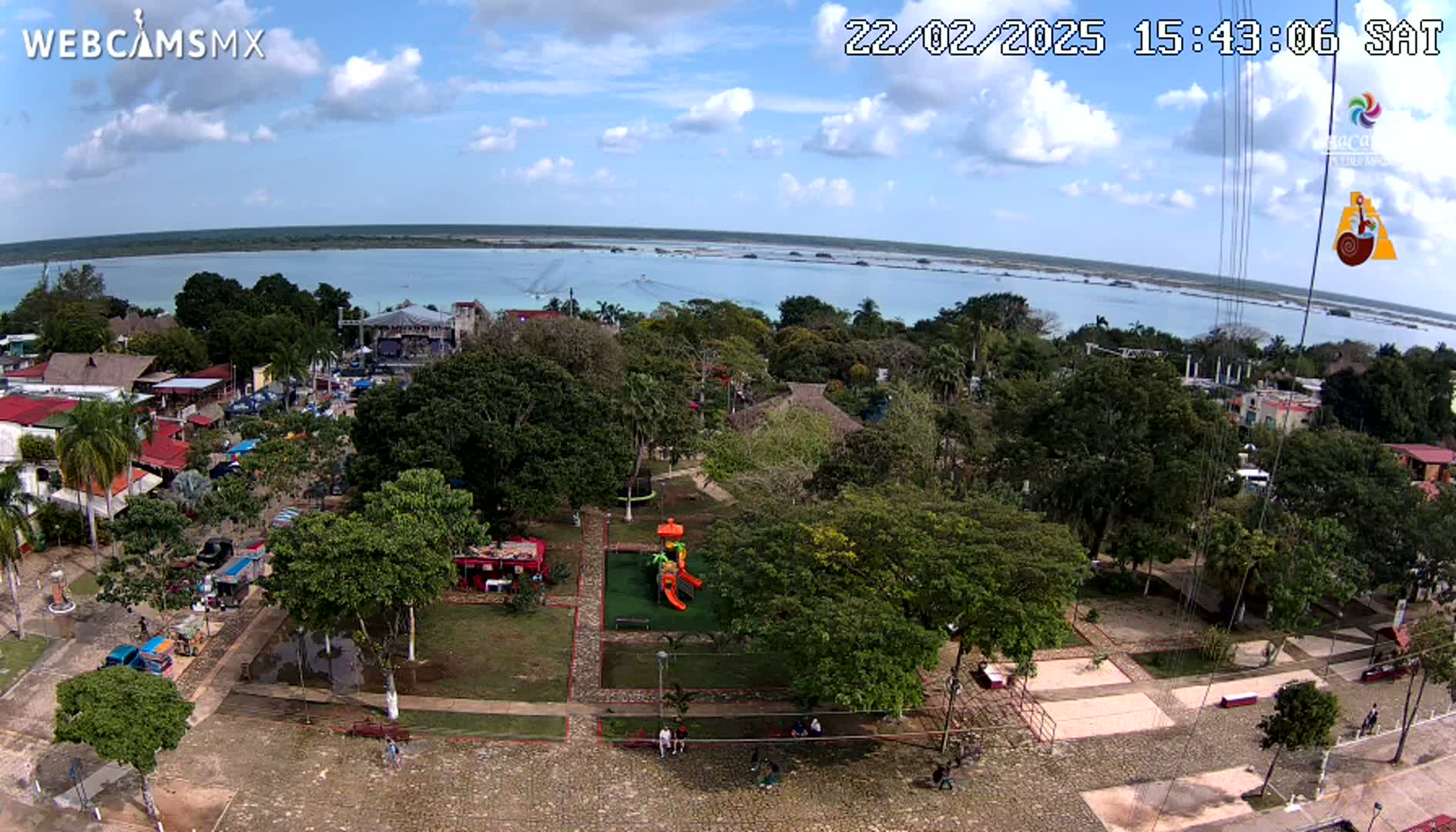 An aerial view of a town square with a playground, lush green trees, and a calm body of water visible in the distance under a partly cloudy sky.