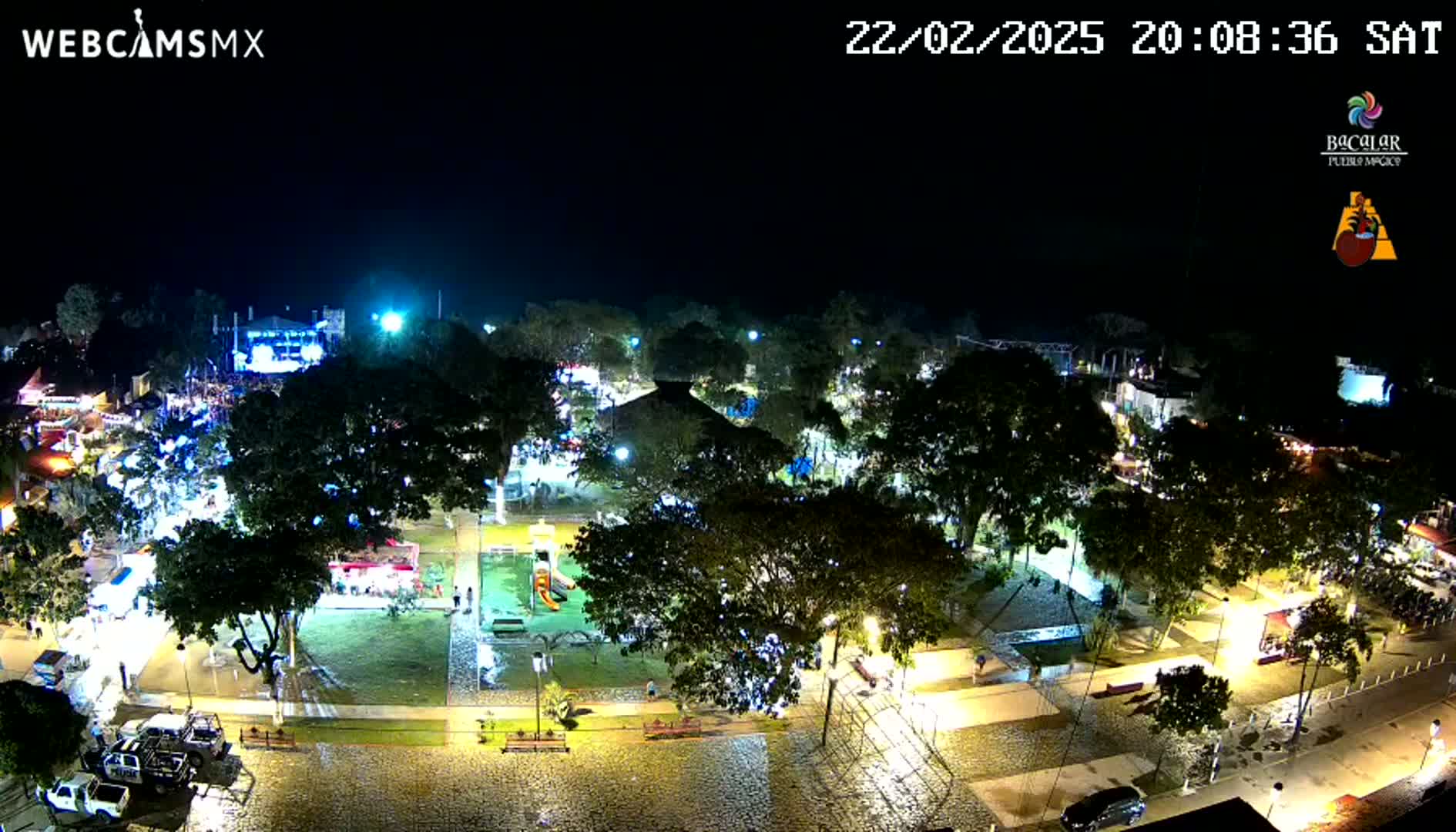 A brightly lit town square at night, surrounded by trees and buildings, under clear skies.