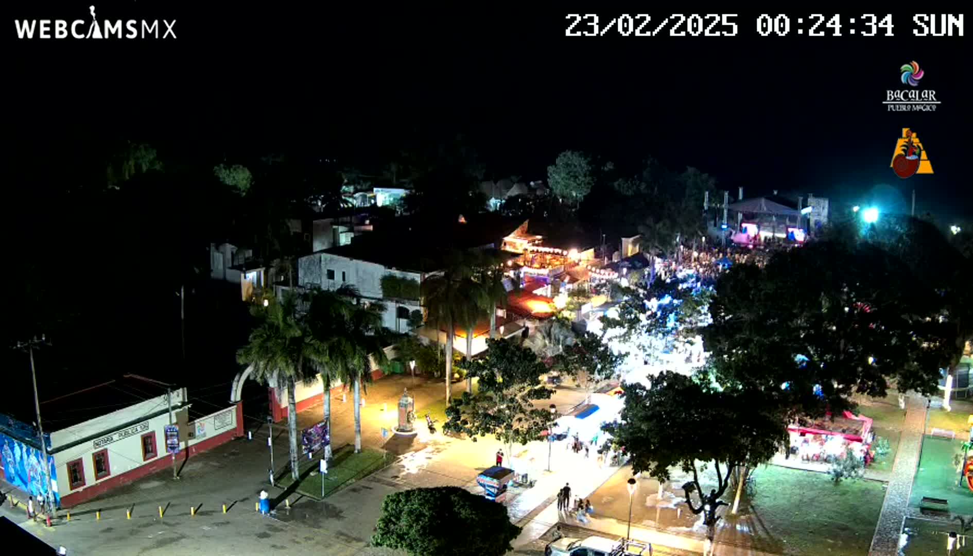A nighttime, aerial view of a town square filled with people and illuminated buildings and stalls under a clear sky.