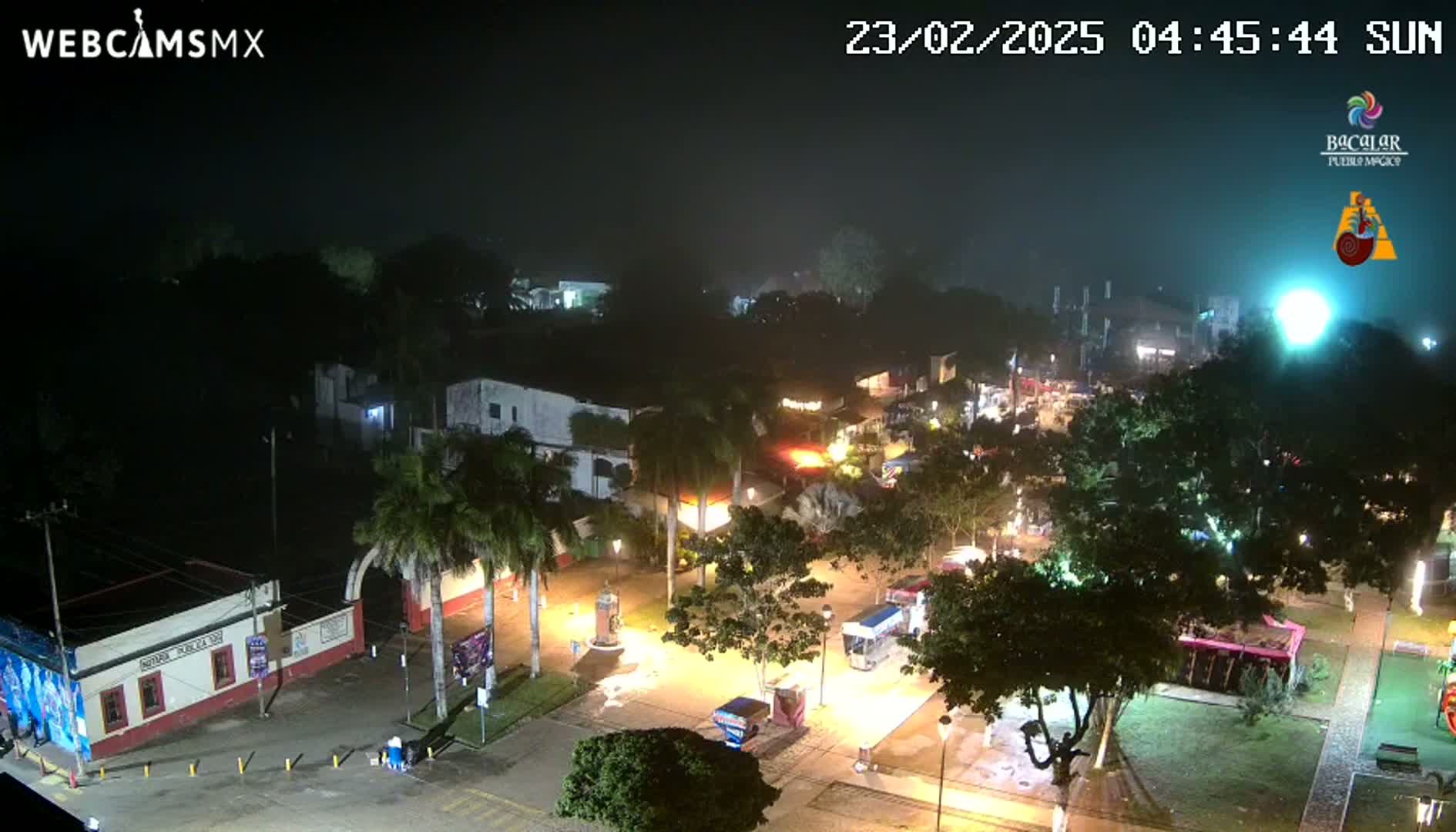 A nighttime, hazy view of a town square with illuminated buildings, trees, and vehicles.