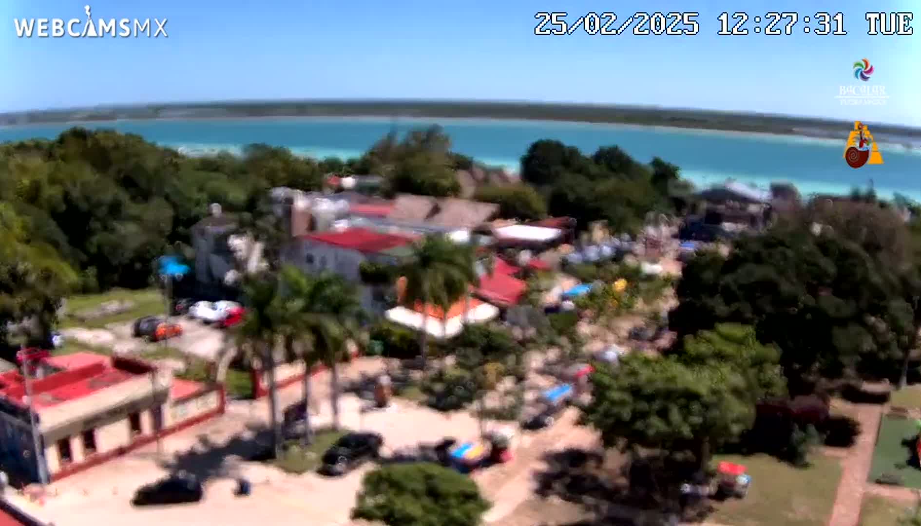 An aerial view shows a town nestled among lush green trees next to a calm, turquoise body of water under a clear, sunny sky.