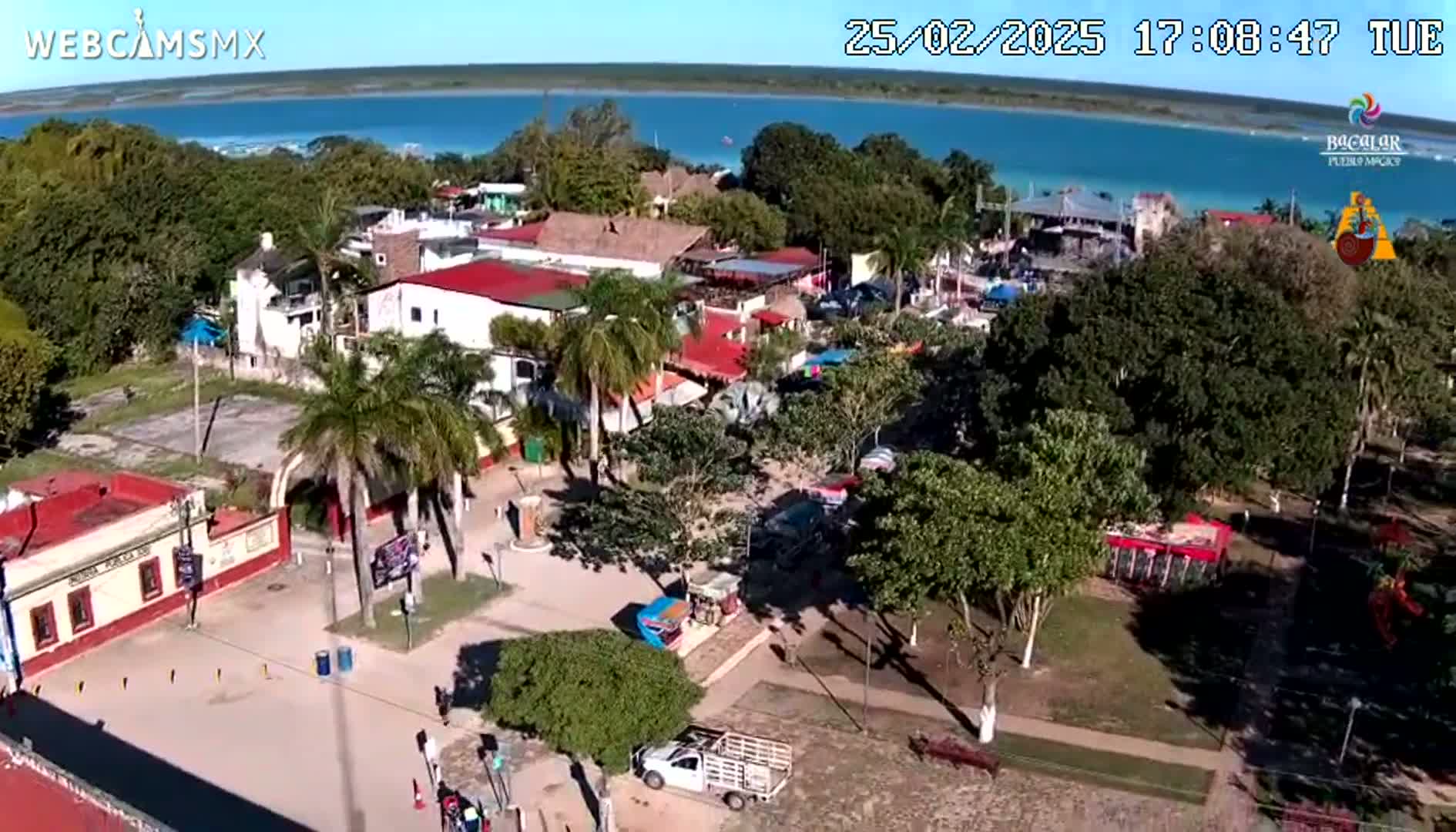 An aerial view shows a town square with buildings, palm trees, and a body of water in the background under sunny skies.