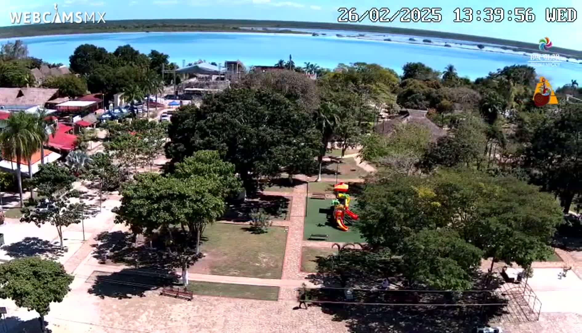 An aerial view shows a town square with a playground, numerous trees, and buildings near a bright blue body of water under sunny skies.