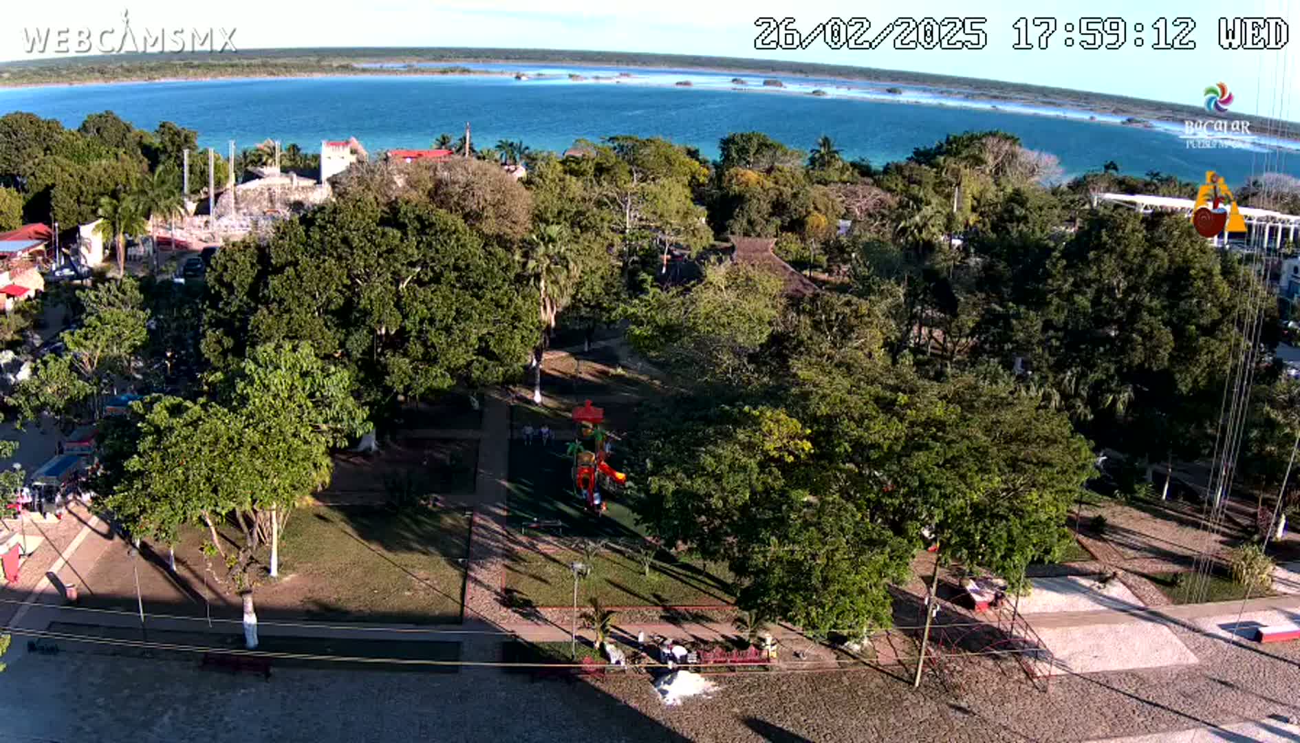 An aerial view shows a park with a playground next to a body of blue water under a sunny sky.