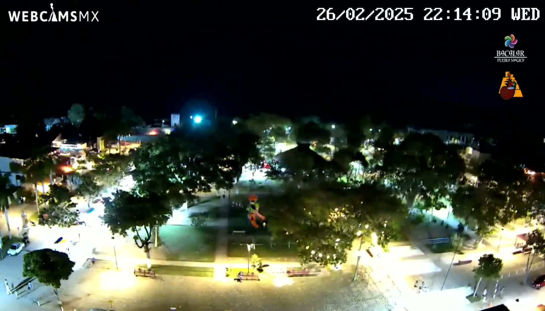 An aerial nighttime view shows a town square illuminated by streetlights, surrounded by trees and buildings.