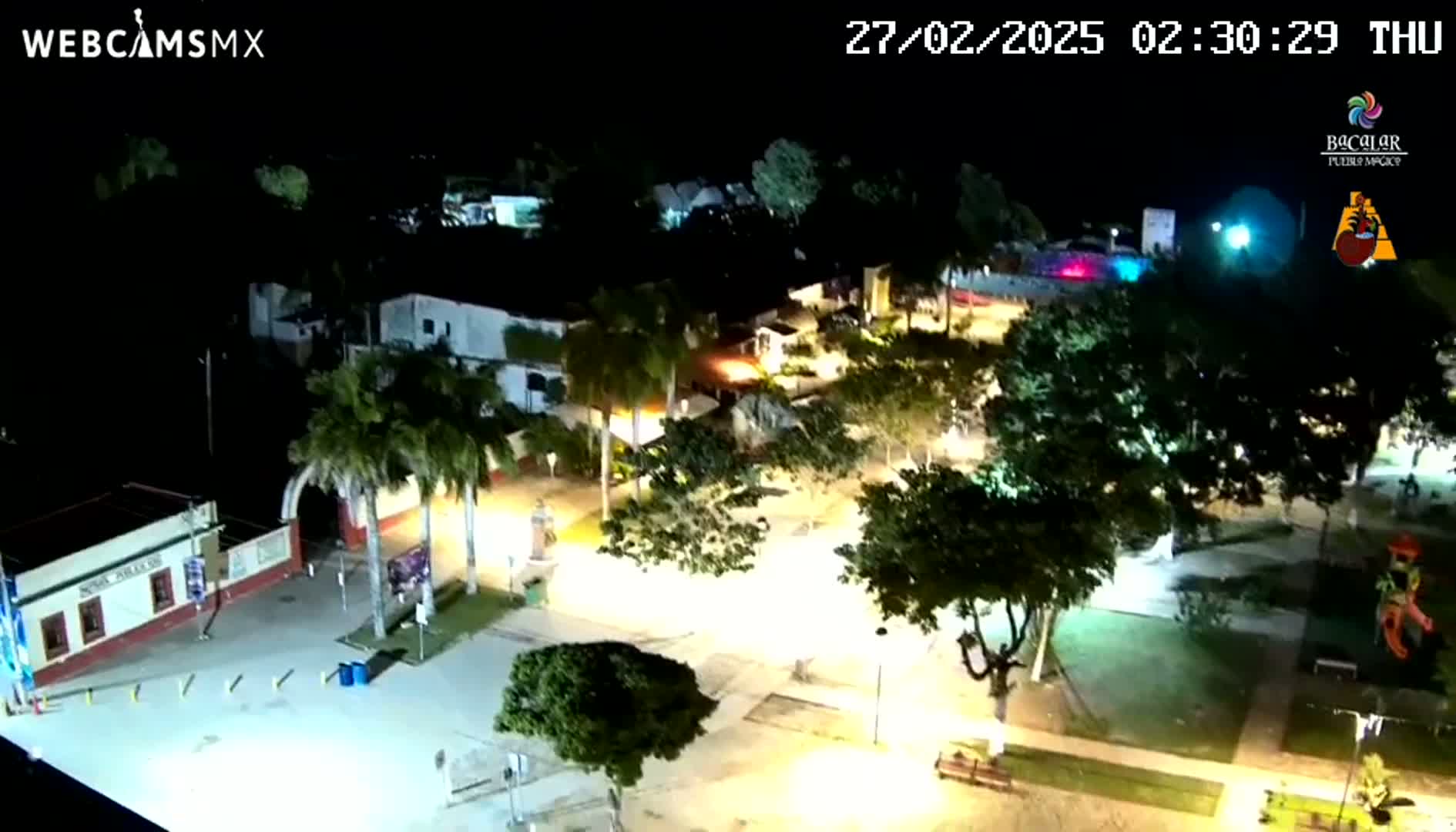 A nighttime, high-angle view of a town square illuminated by streetlights, featuring palm trees, buildings, and a few people.