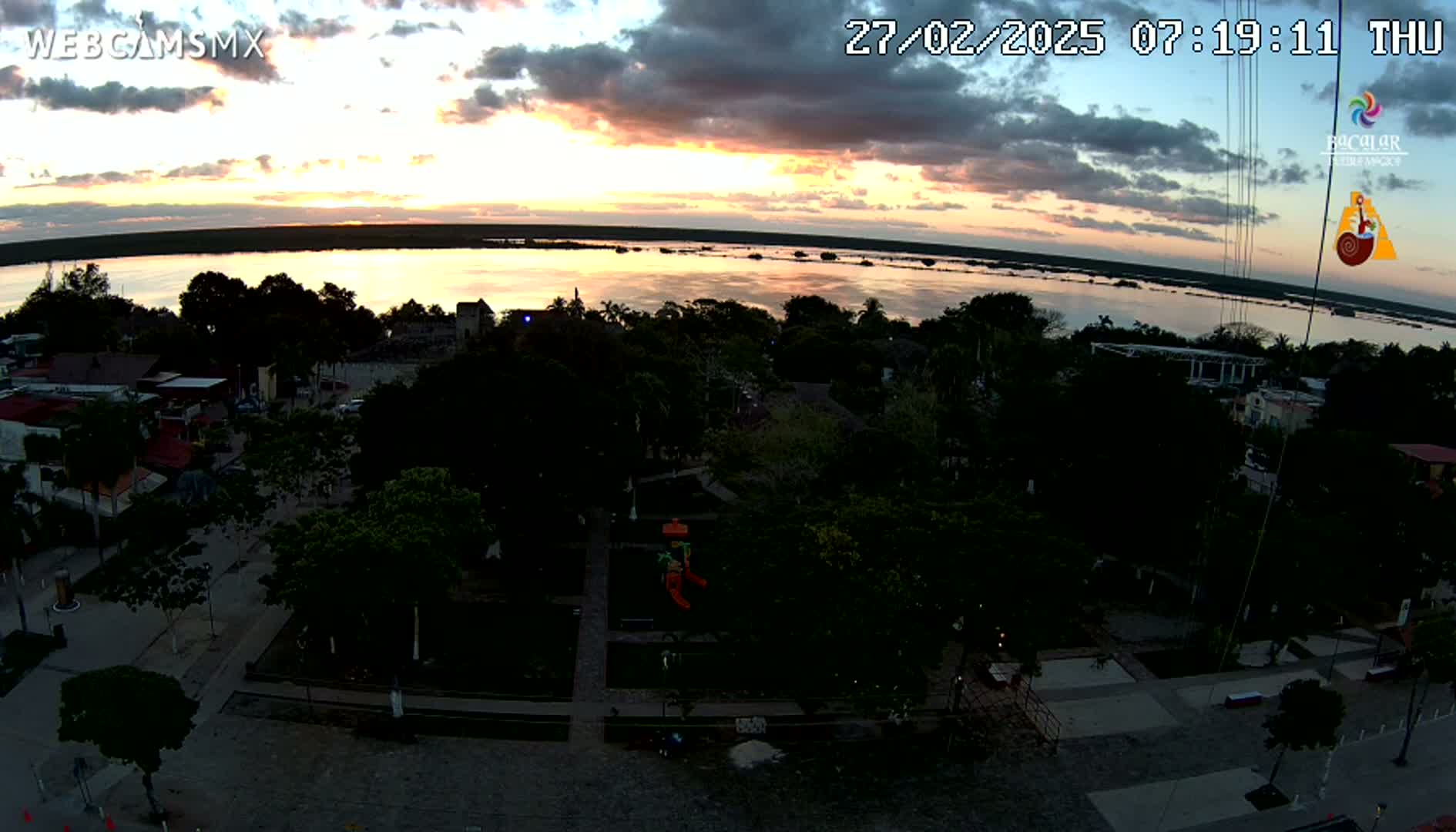 An aerial view at sunset shows a town next to a calm body of water, with a playground visible in a central park.