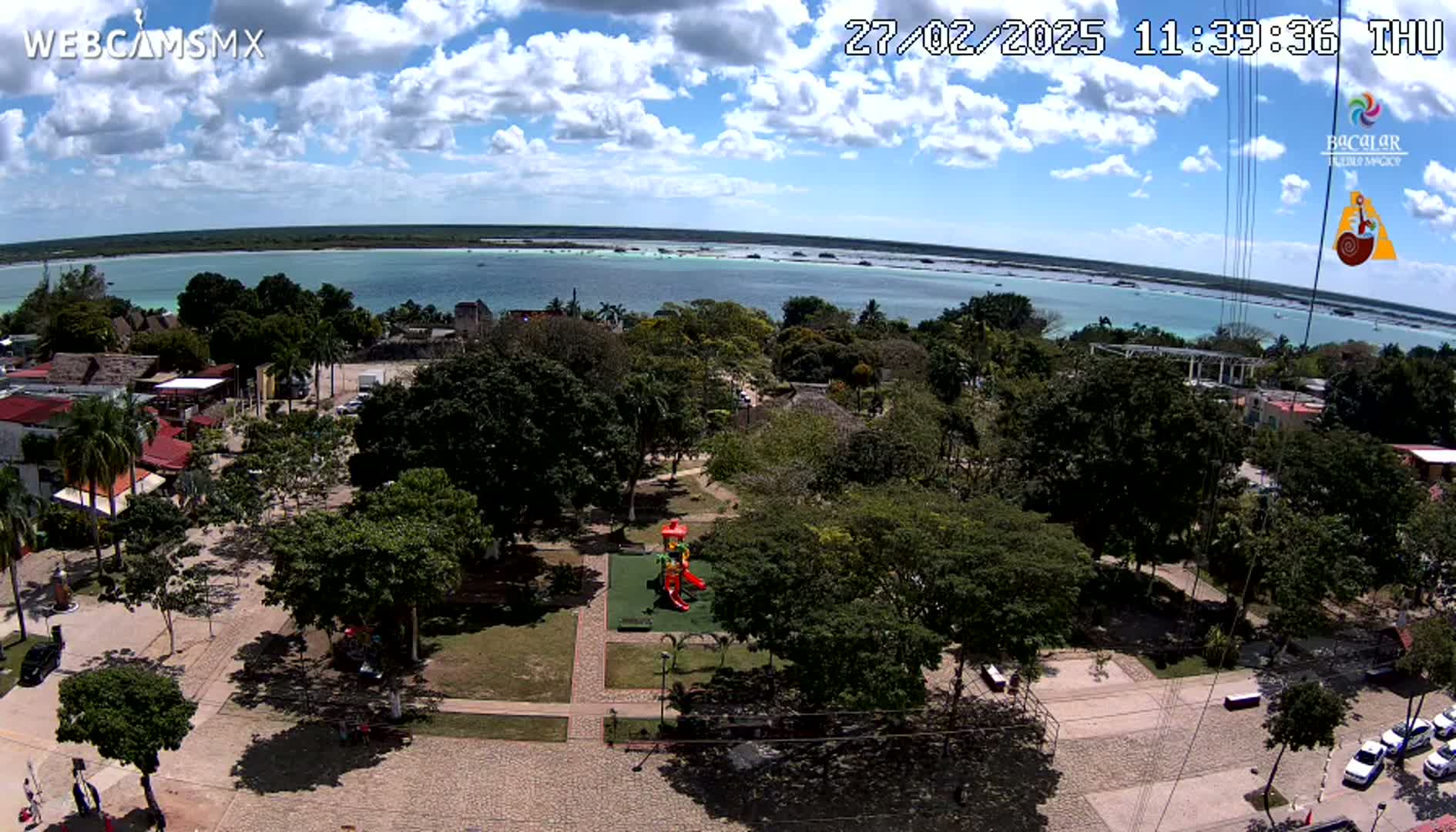 A partly sunny aerial view of a town square with a playground, surrounded by lush green trees and overlooking a calm body of water under a partly cloudy sky.