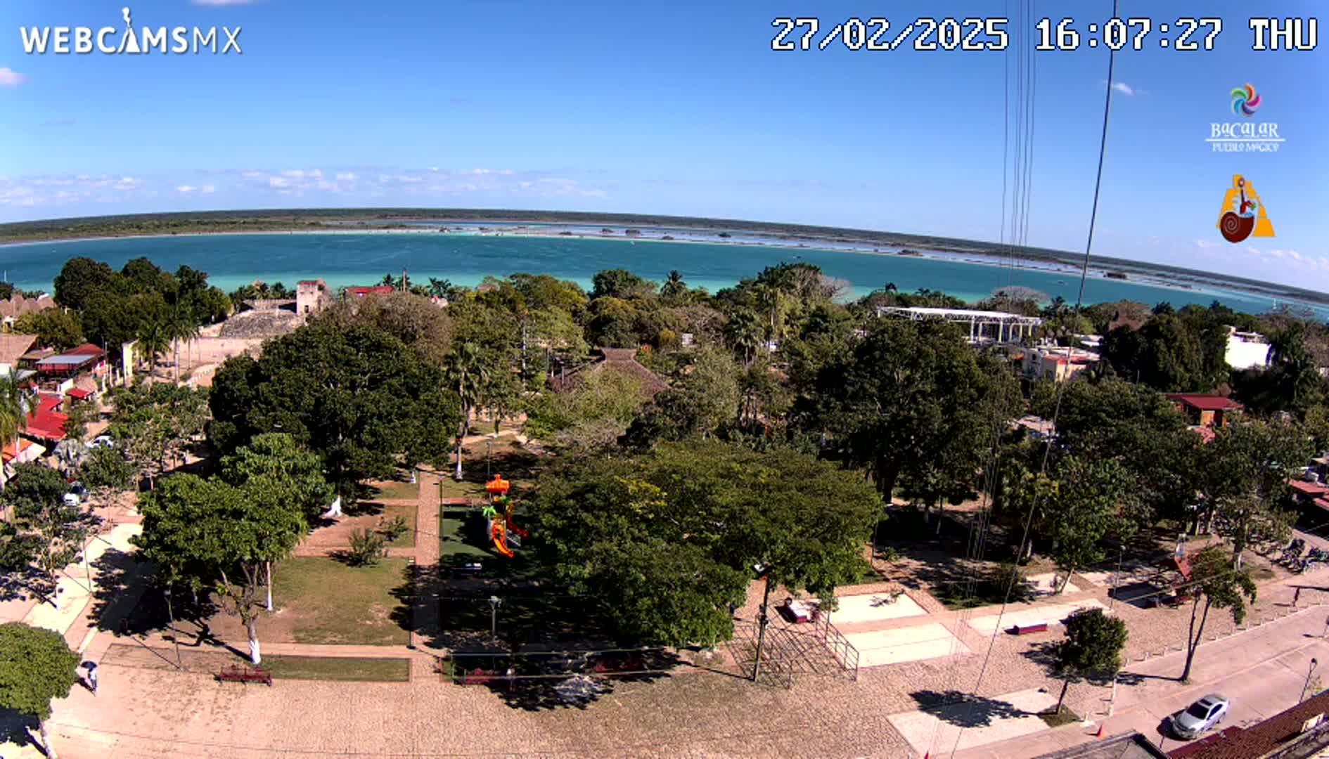 An aerial view shows a town square with lush green trees and a playground next to a calm, turquoise body of water under a clear blue sky.