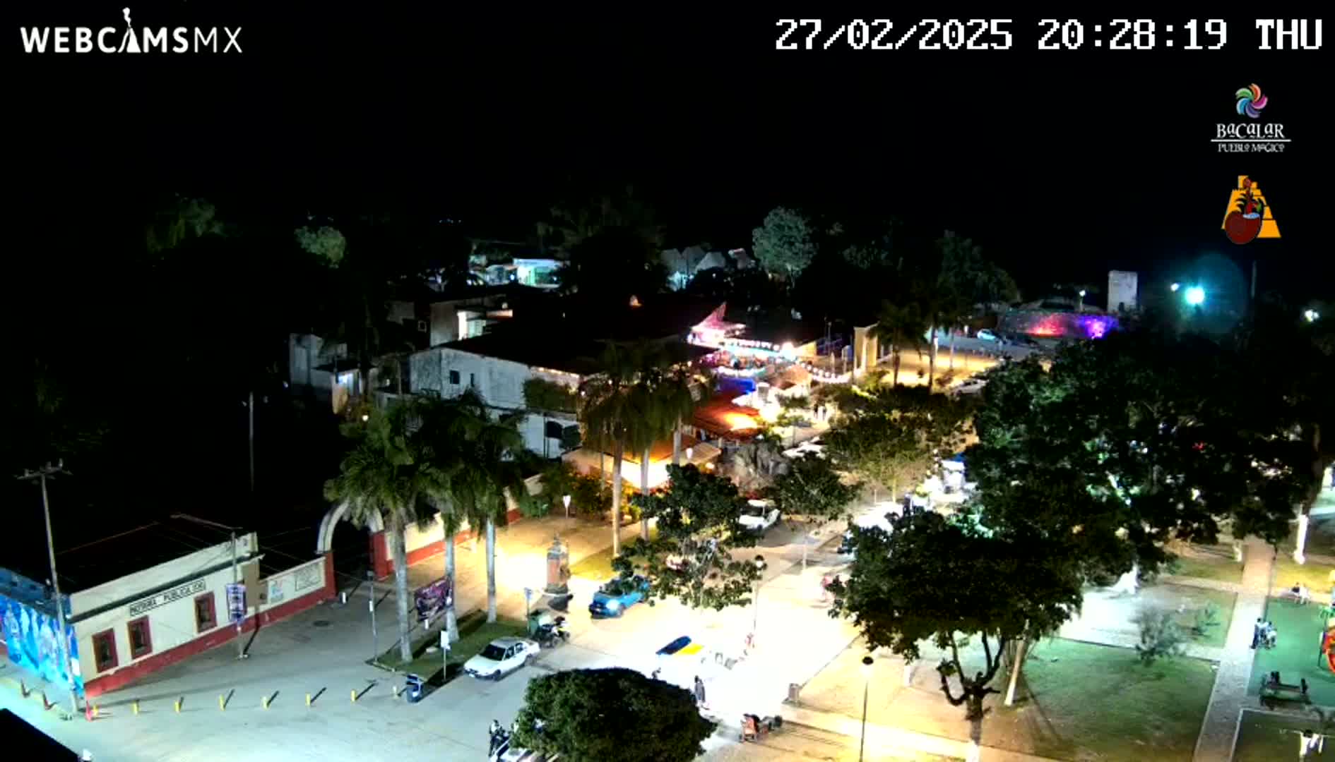 A nighttime aerial view of a town square, illuminated by streetlights, showing buildings, parked cars, and trees under a clear sky.