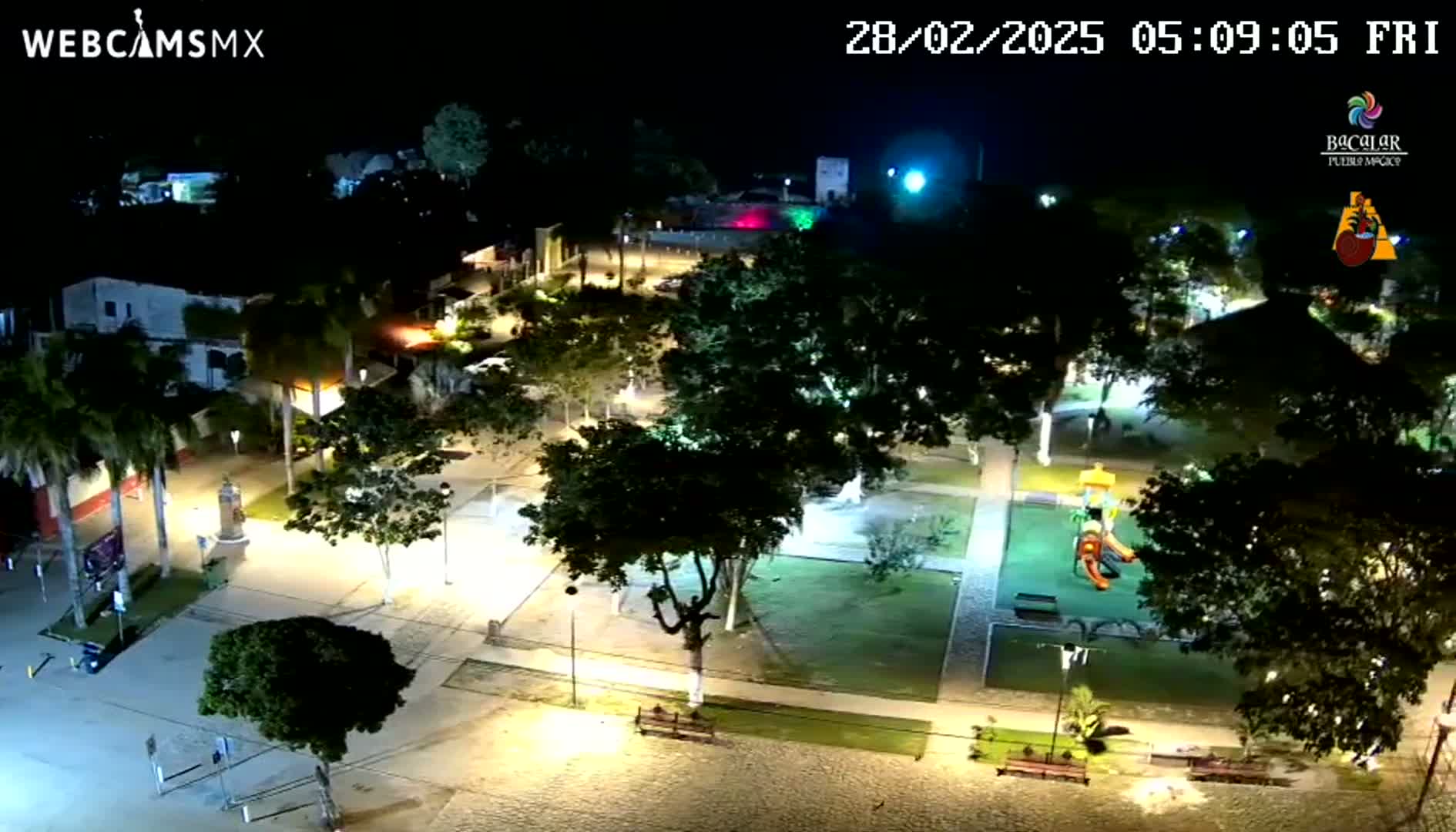 A nighttime aerial view of a town square with illuminated pathways, trees, and a playground.