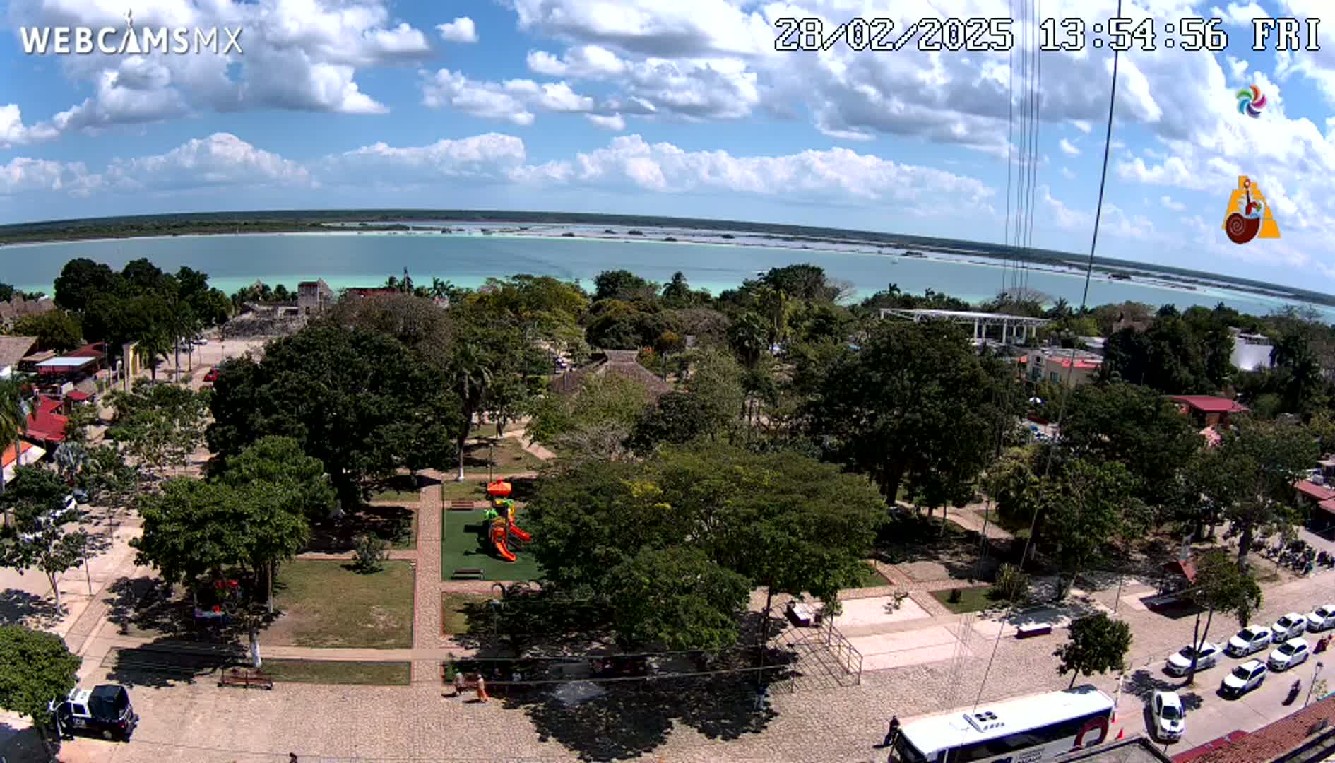 An aerial view shows a town square with a playground, lush green trees, and parked cars, next to a calm, light-blue body of water under a partly cloudy sky.