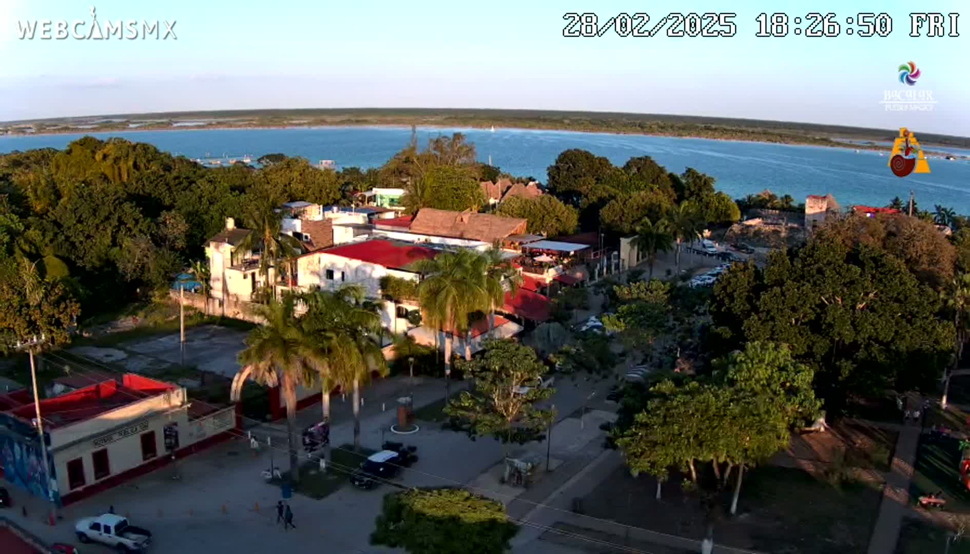 An aerial view shows a town with buildings near a calm body of water under a clear, sunset sky.