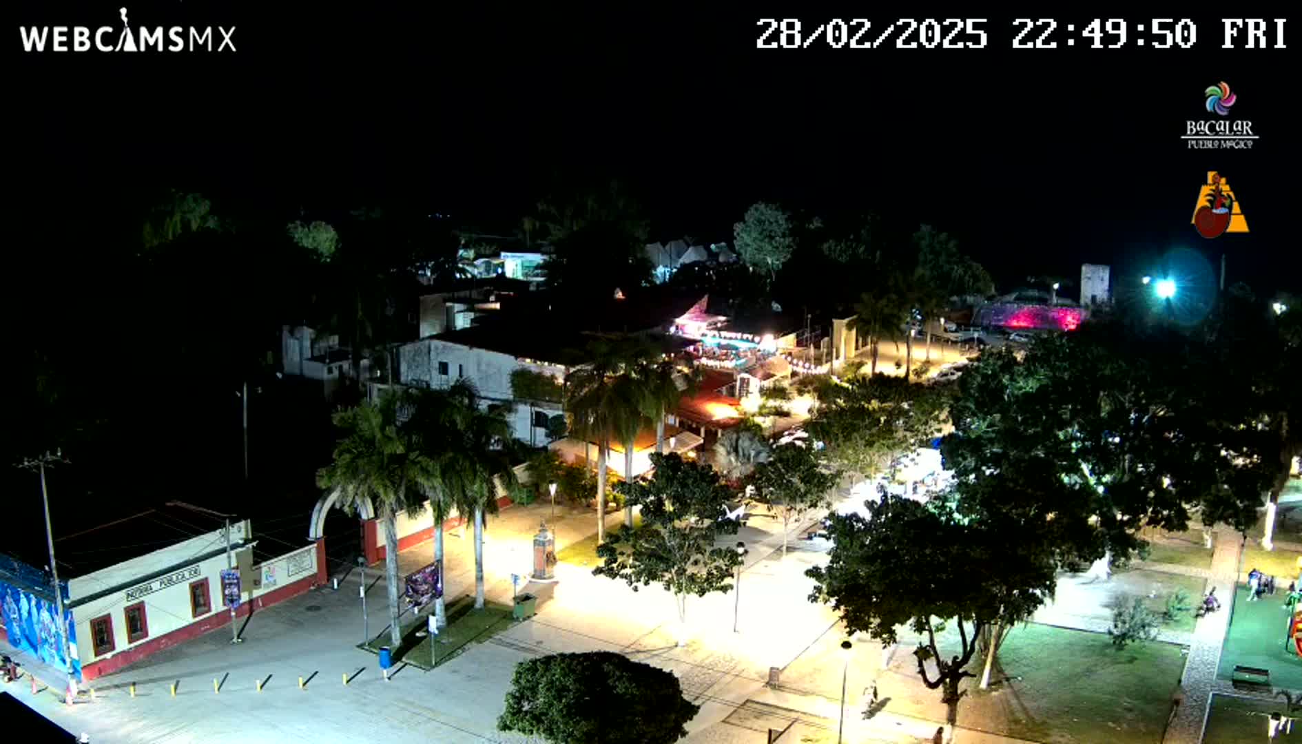 An aerial nighttime view of a town square with palm trees, buildings, and lights, under a clear, dark sky.
