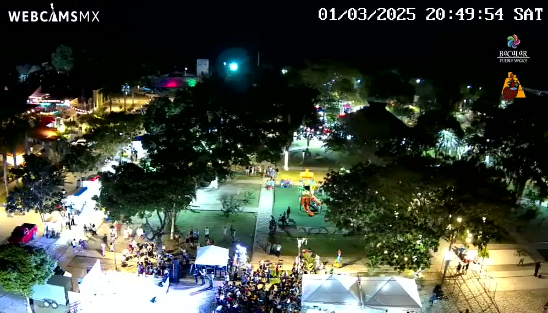 A nighttime aerial view shows a town square with numerous people gathered around tents and a stage, surrounded by lush, illuminated trees under a clear sky.