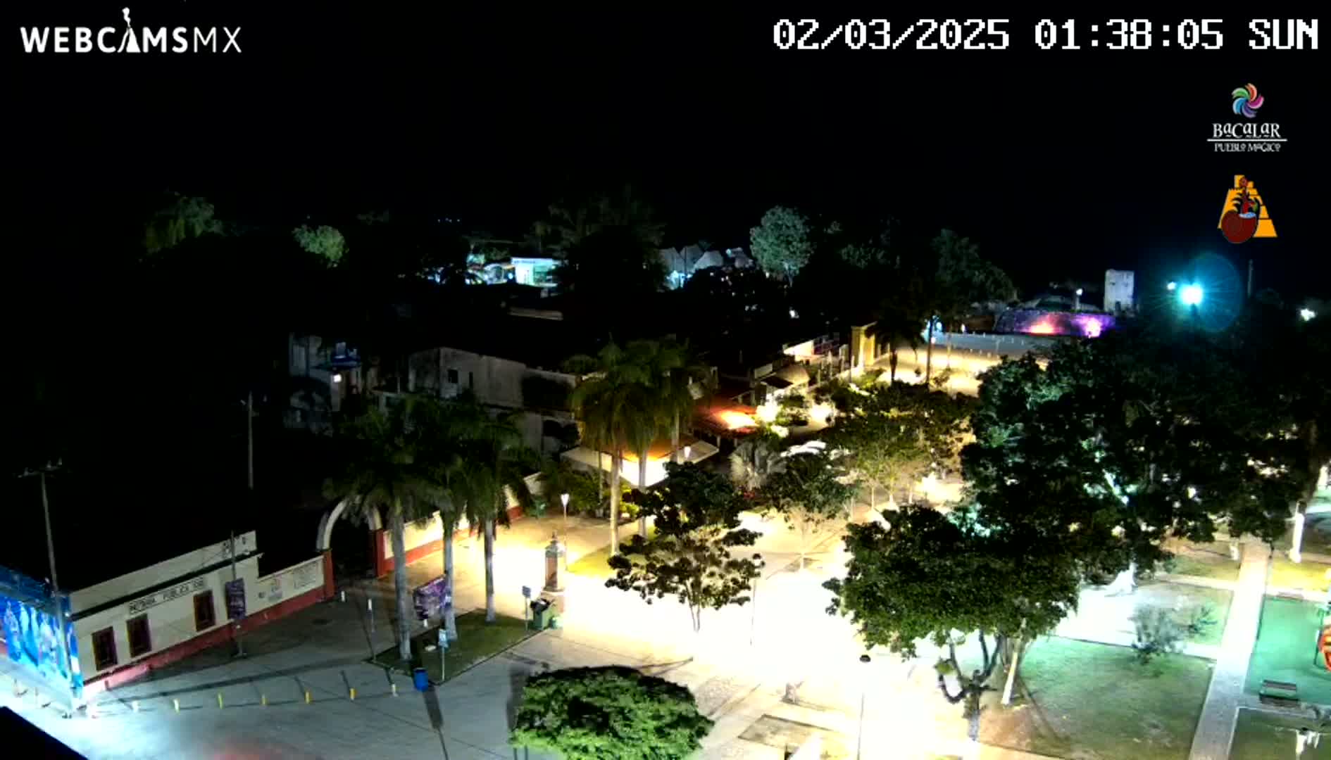 A nighttime, overhead view of a town square, illuminated by streetlights, featuring palm trees and buildings.