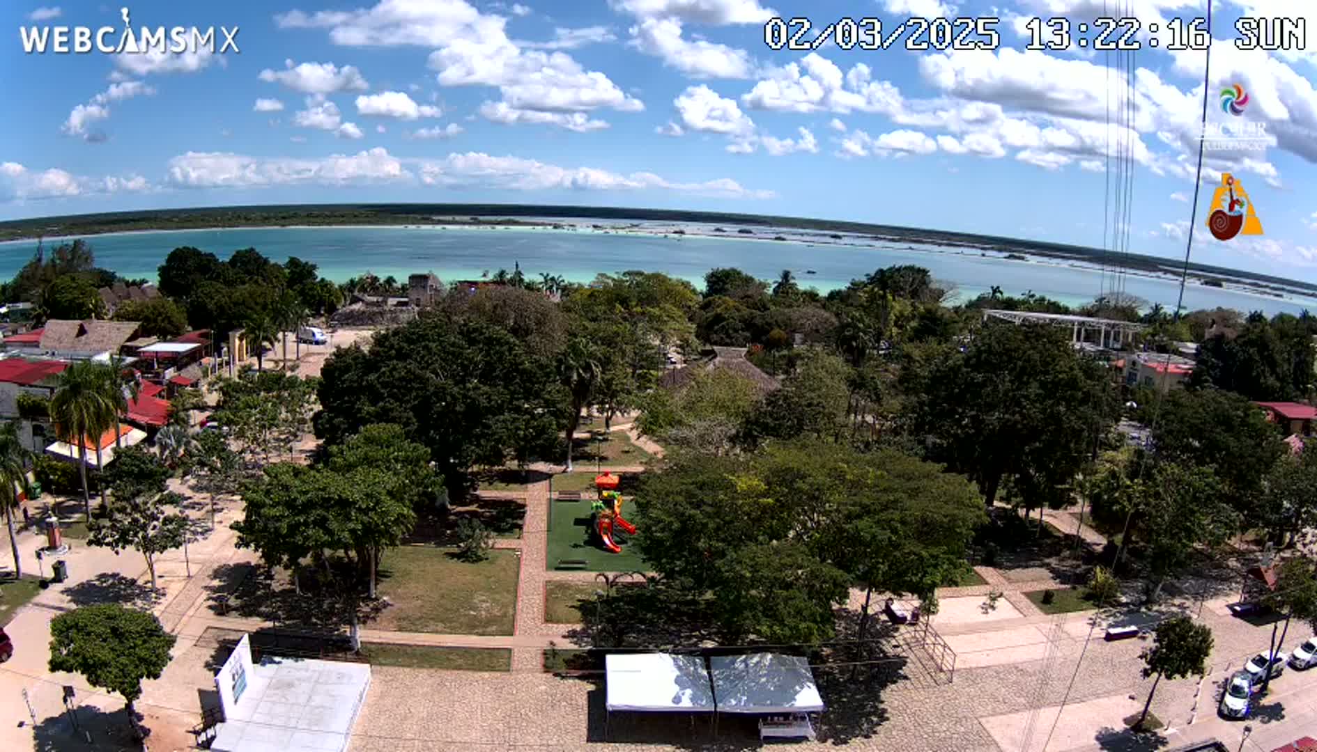 An aerial view of a town square with a playground, surrounded by lush green trees, near a calm, light-blue body of water under a partly sunny sky.