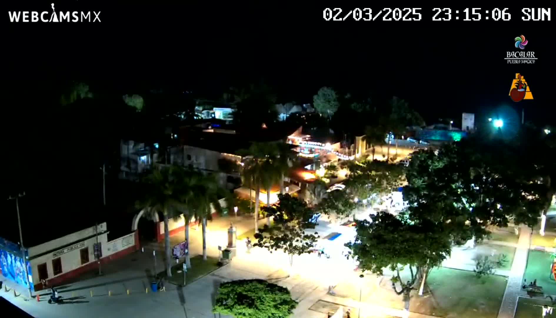 An aerial nighttime view of a town square with palm trees and buildings illuminated, under a clear sky.