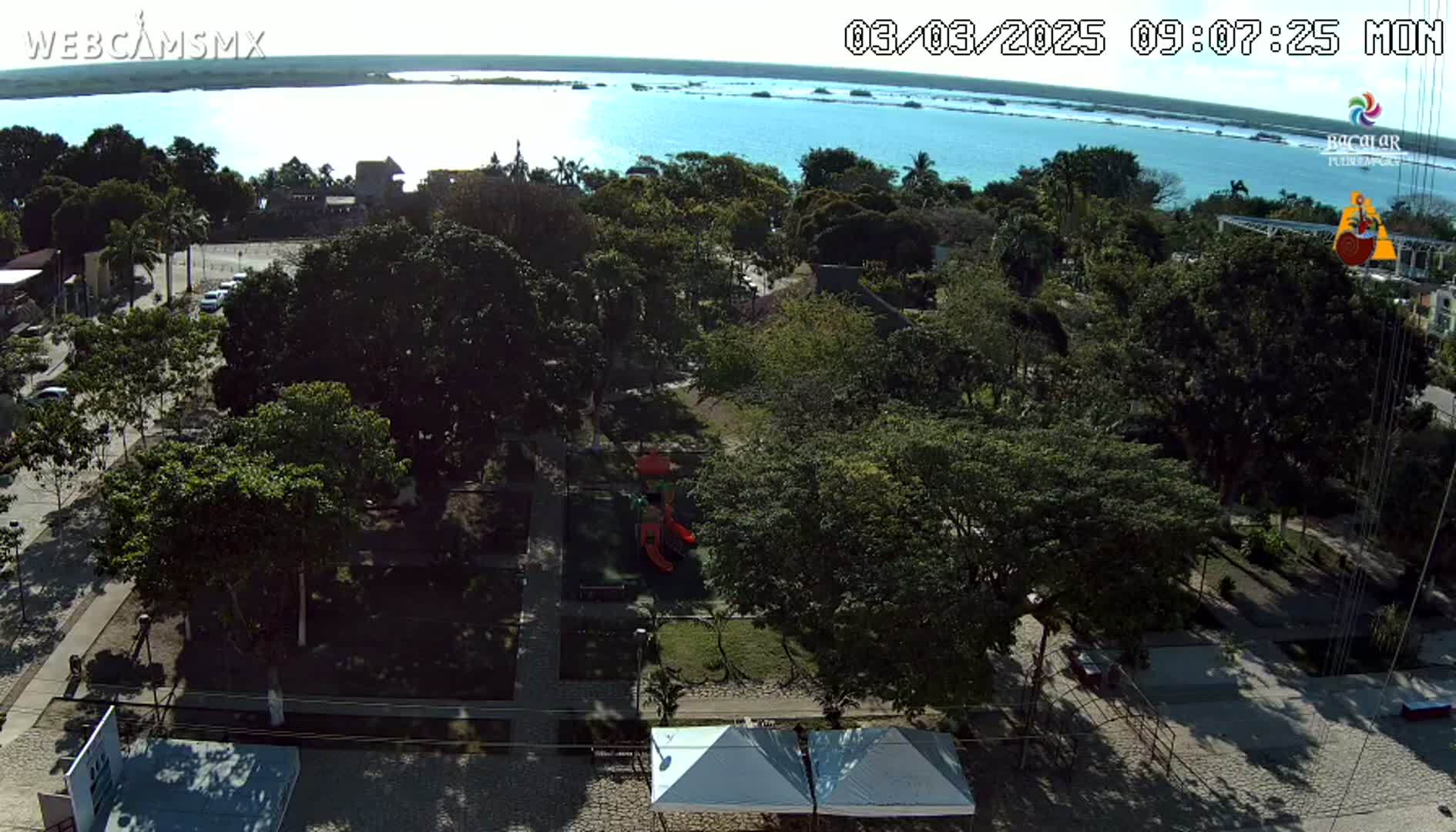 A sunny day shows a park with playground equipment and several leafy trees next to a calm body of water.