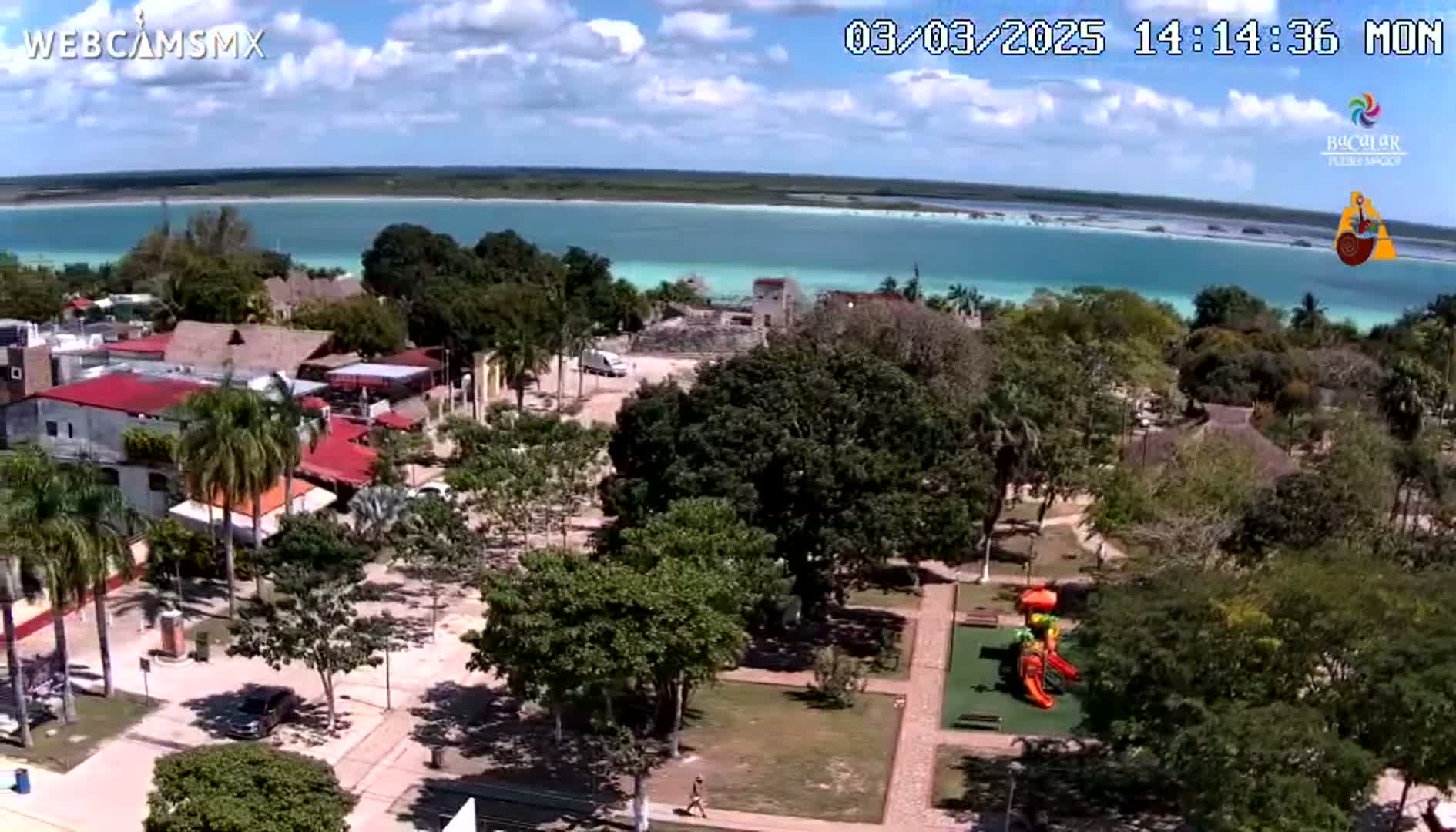 An aerial view of a town next to a bright turquoise body of water on a partly sunny day shows a park with a playground in the foreground.