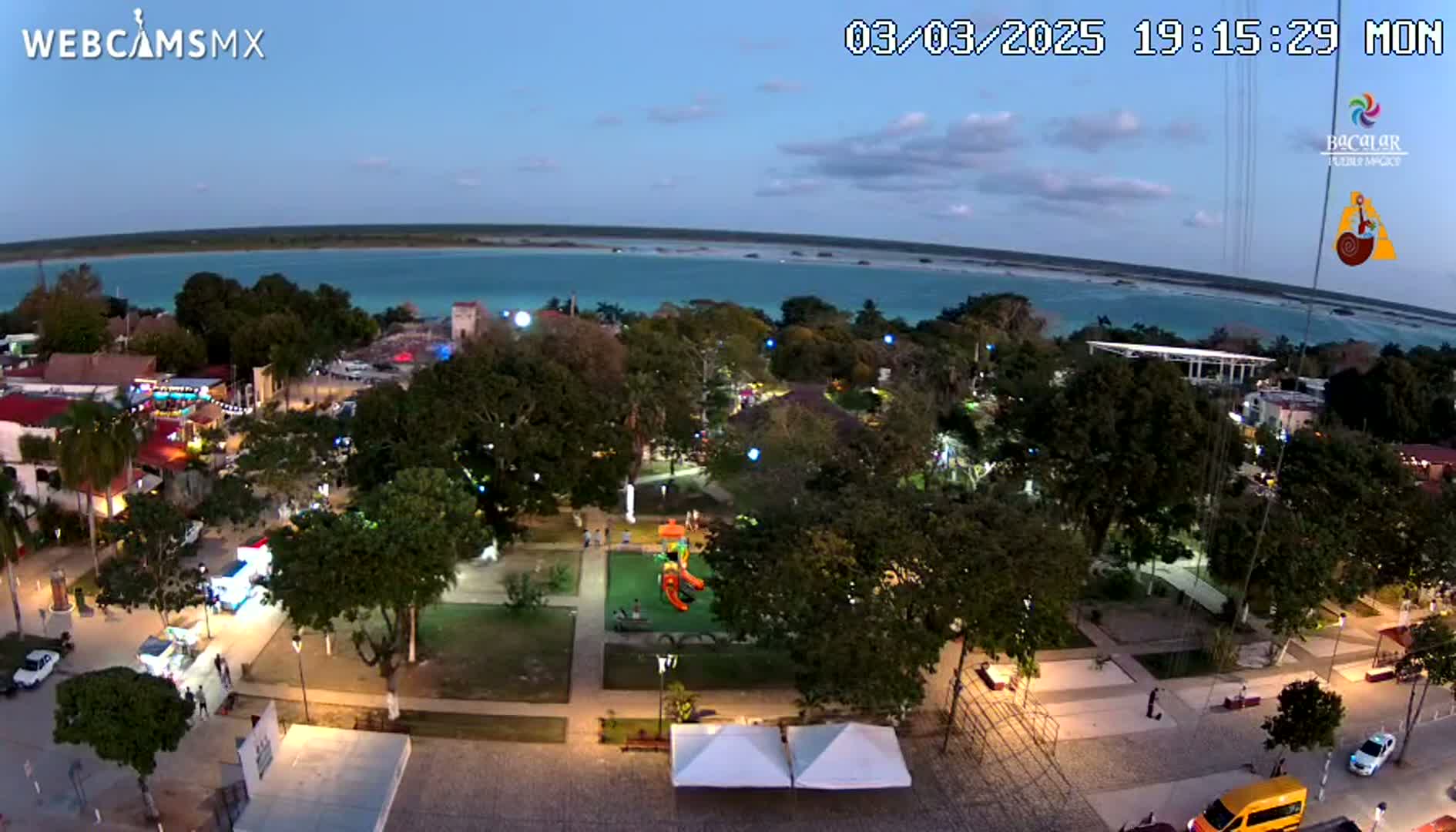 An aerial evening view of a town square with a playground, illuminated streets, and lush greenery, overlooking a calm turquoise body of water under a twilight sky.