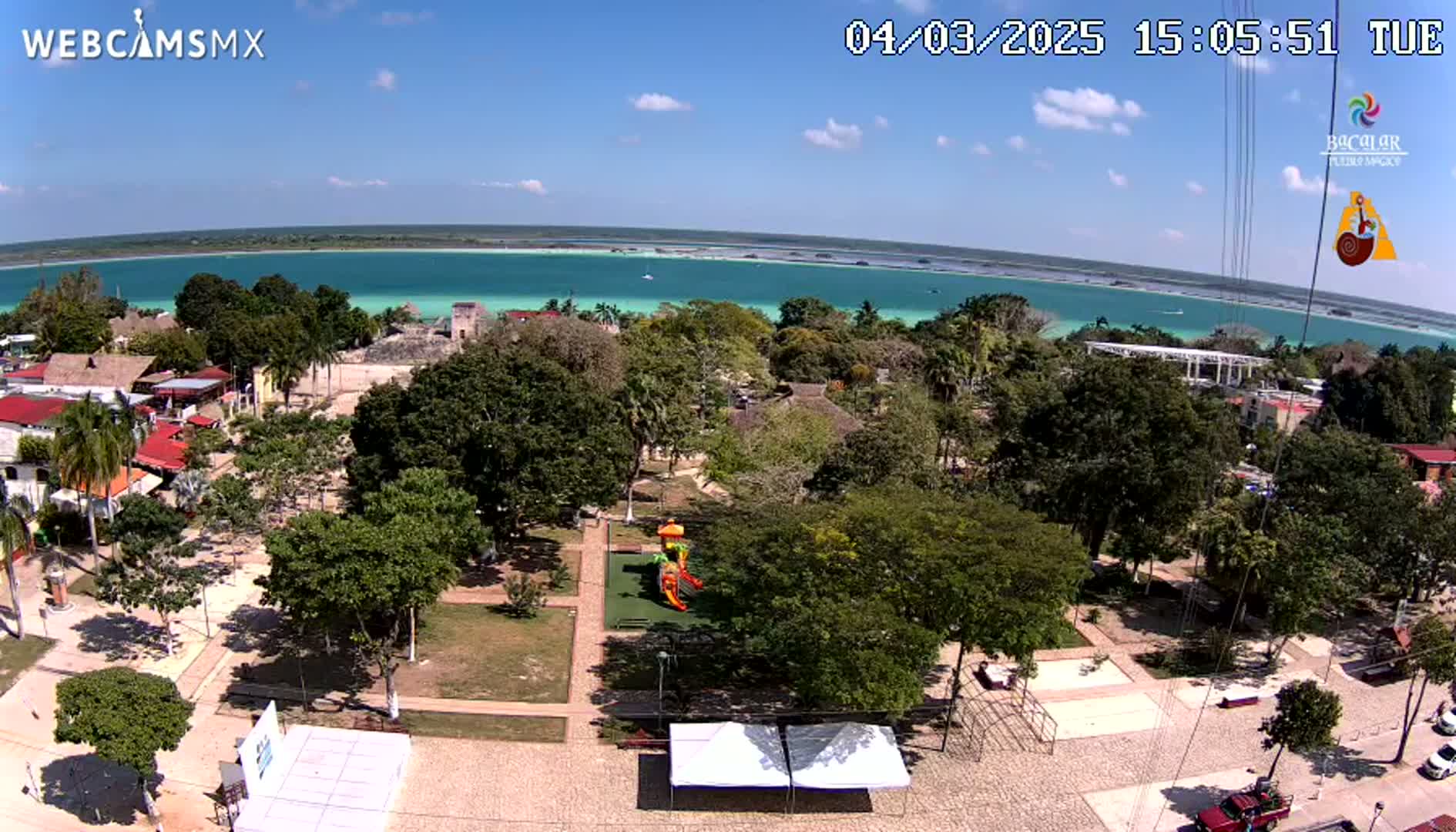 An aerial view shows a town square with a playground next to a park, all overlooking a bright turquoise body of water under a mostly sunny sky.
