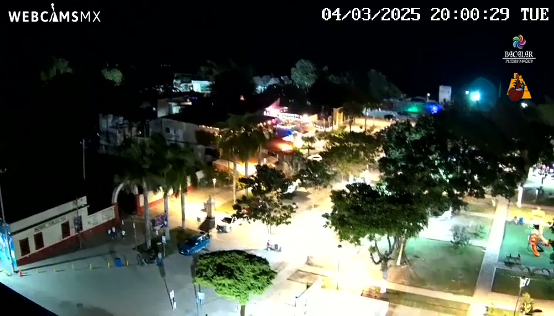 An illuminated town square at night, showing buildings, trees, and a few vehicles, under clear skies.