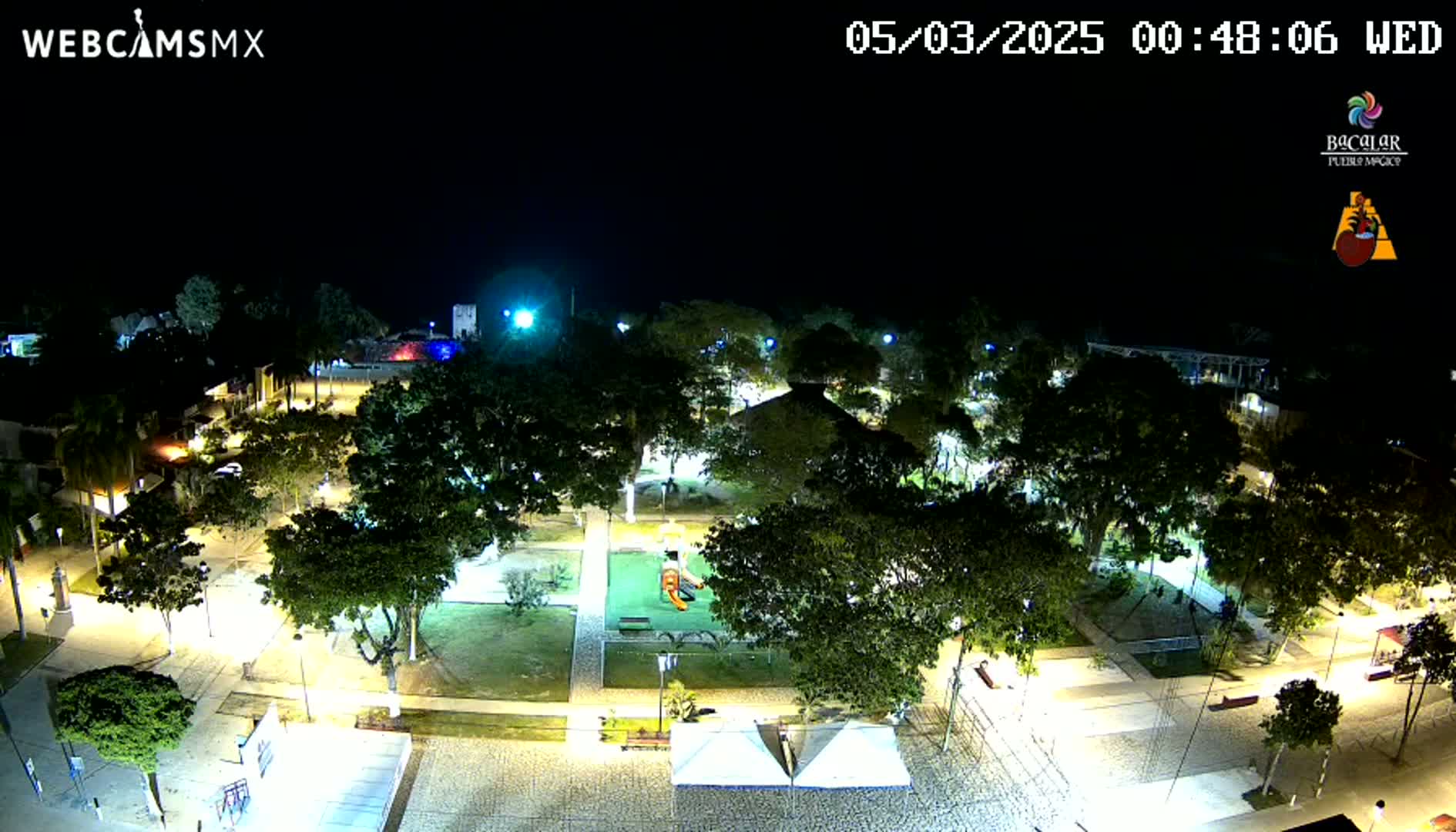 A brightly lit plaza at night, filled with lush green trees and a playground, under a clear, dark sky.
