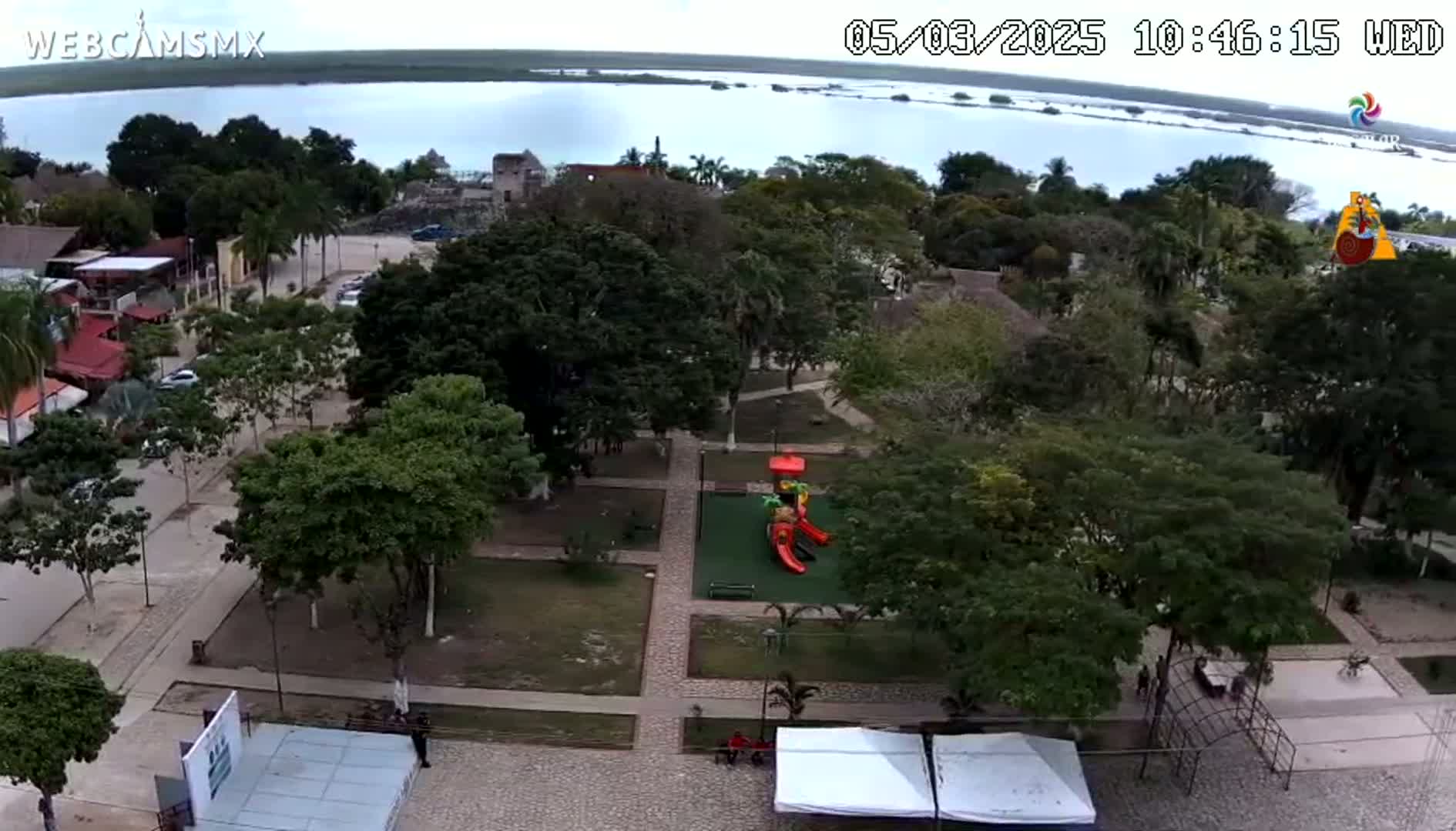 An aerial view shows a town square with a playground, surrounded by lush green trees and buildings, under a mostly cloudy sky overlooking a body of water.
