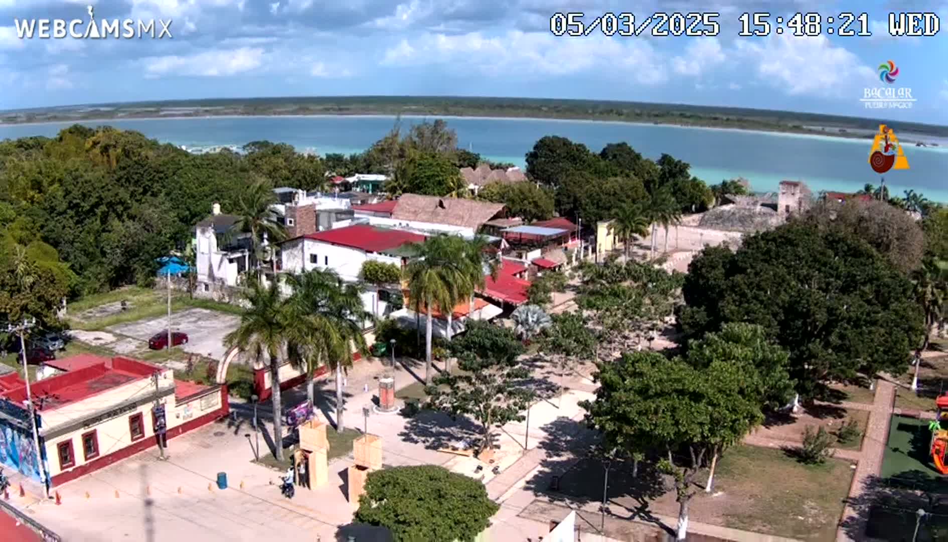 An aerial view shows a town square with palm trees, buildings with red roofs, and a large body of water in the background under a partly cloudy sky.