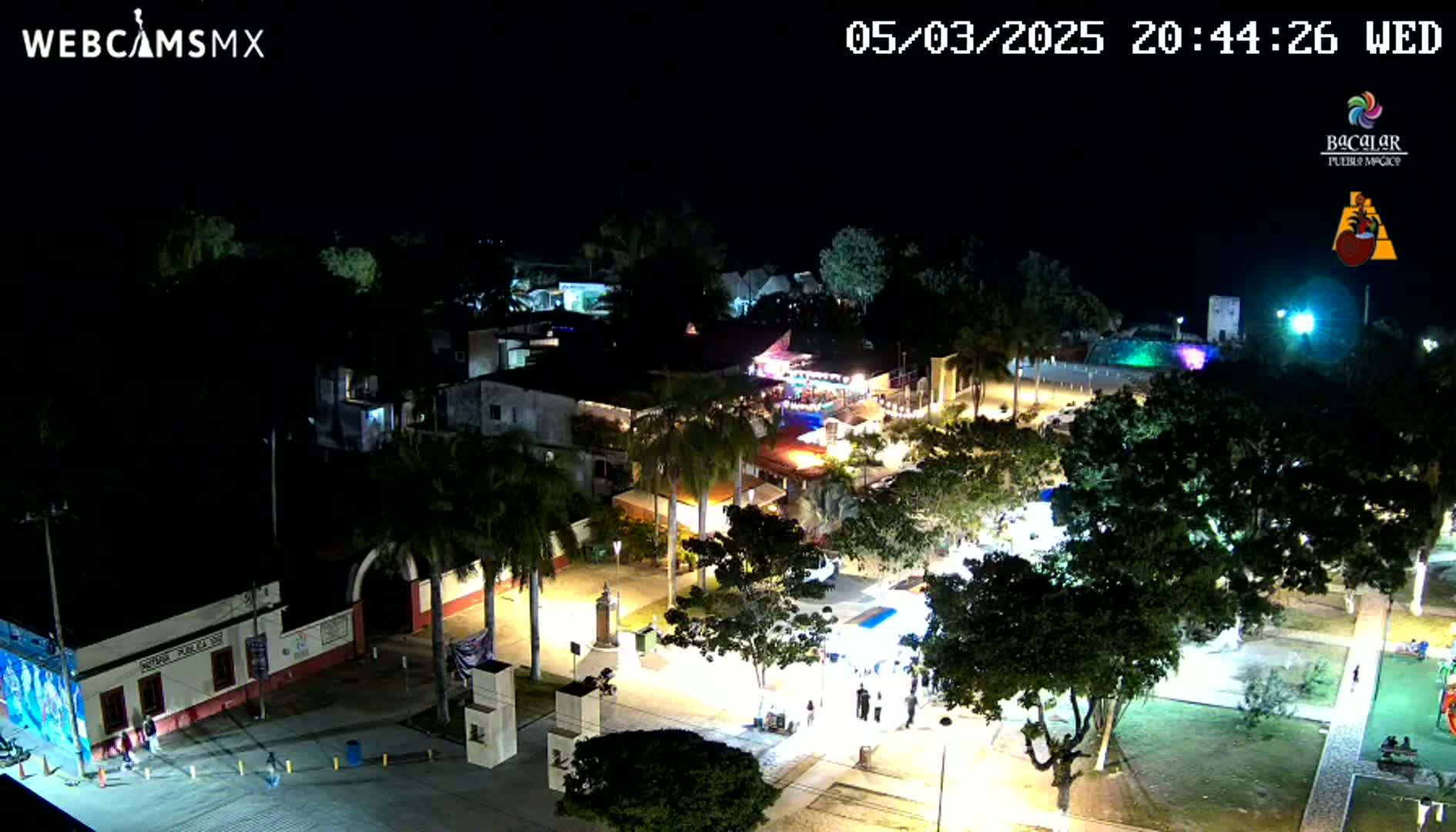 A nighttime aerial view of a town square with palm trees, buildings, and people, illuminated by streetlights under a clear, dark sky.