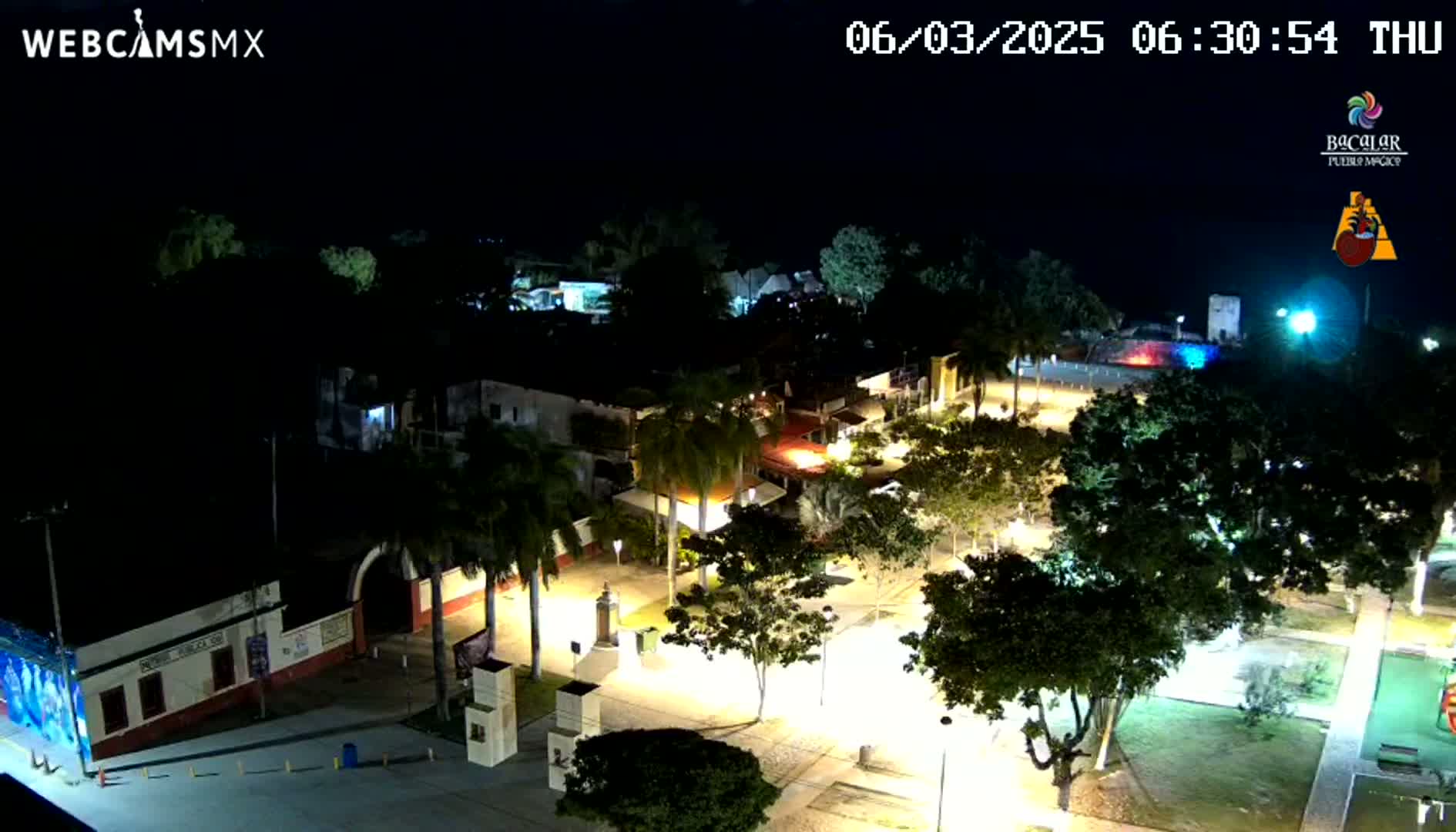 An aerial nighttime view of a town square, illuminated by streetlights, with palm trees and buildings surrounding it under a clear sky.