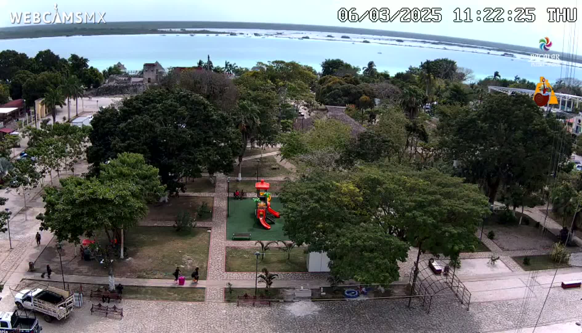 An overhead view shows a park with a playground, surrounded by lush green trees and stone pathways, overlooking a calm, light-blue body of water under a partly cloudy sky.