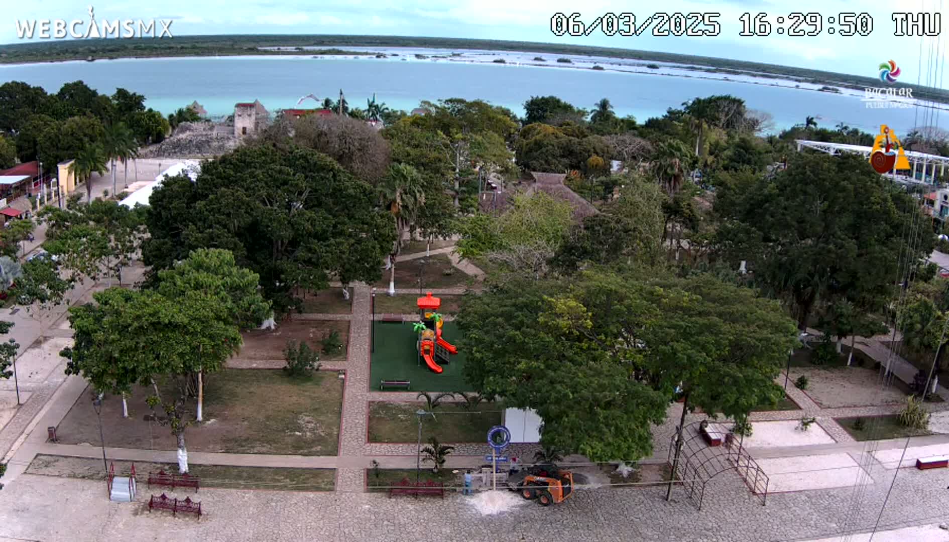 An aerial view of a town square with a playground, surrounded by lush green trees and paved pathways, overlooks a calm body of turquoise water under a partly cloudy sky.