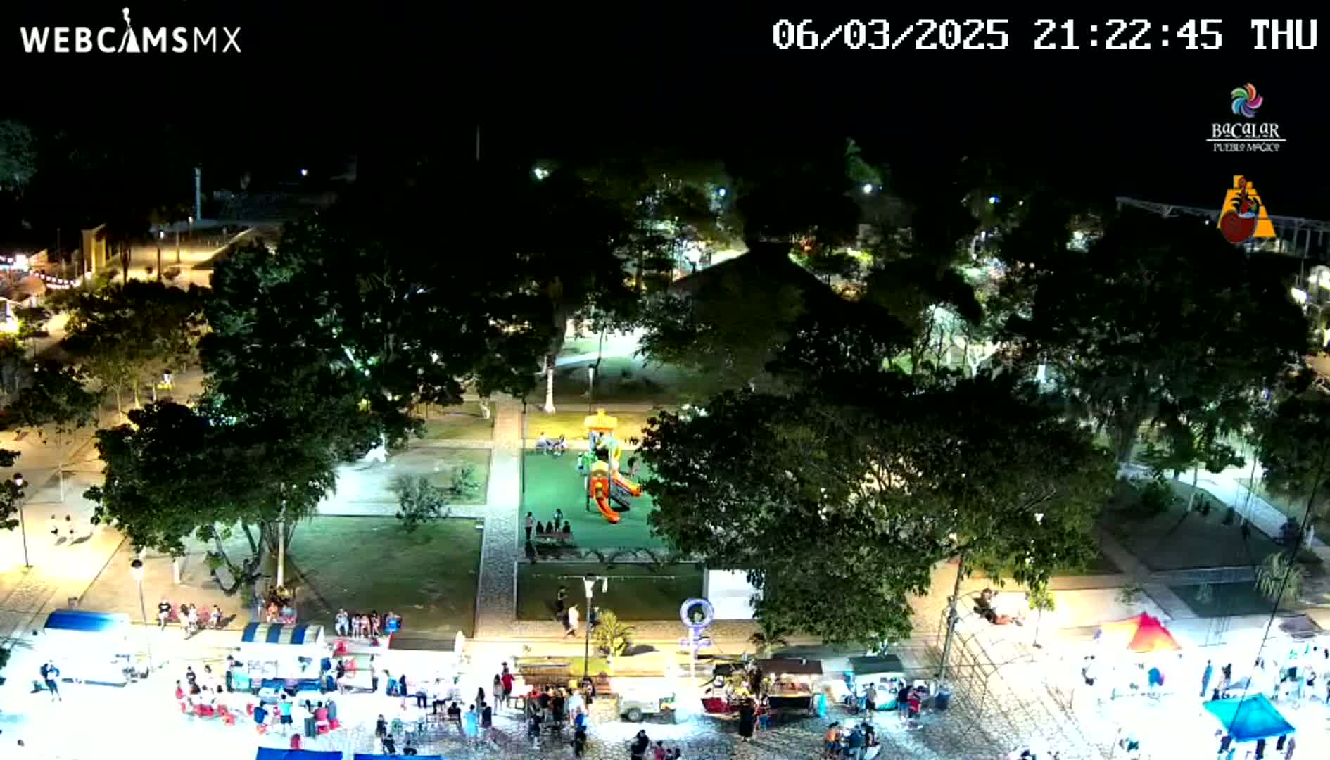 A nighttime aerial view shows a town square with a playground, food vendors, and people gathered under the lights.