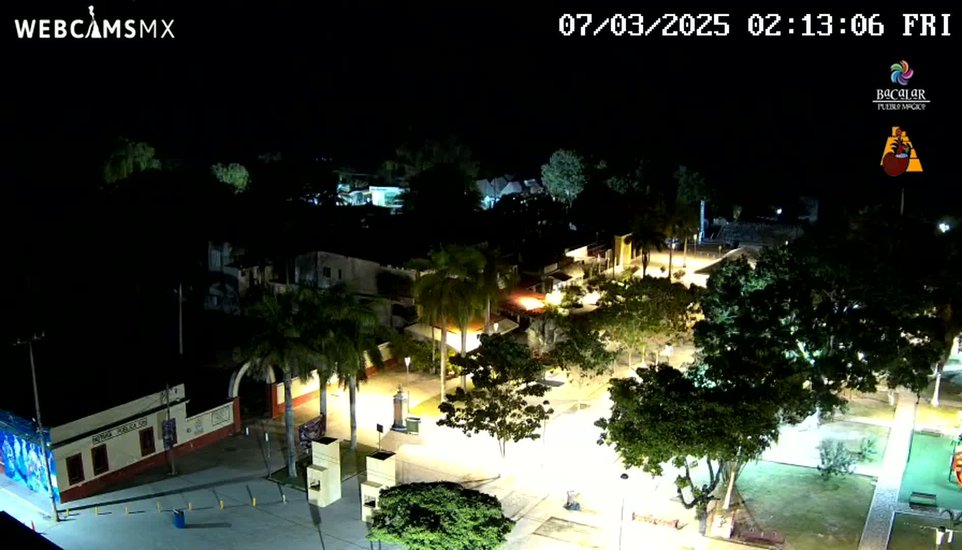 An aerial nighttime view of a town square, illuminated by streetlights, featuring palm trees and buildings.