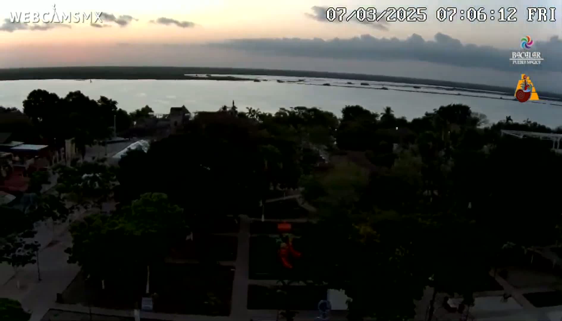 An elevated view shows a town at twilight next to a calm body of water under a mostly cloudy sky.