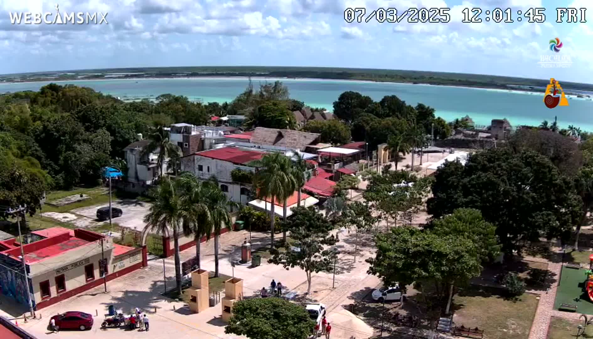 An aerial view shows a town square with palm trees and buildings near a calm, turquoise body of water under a partly cloudy sky.