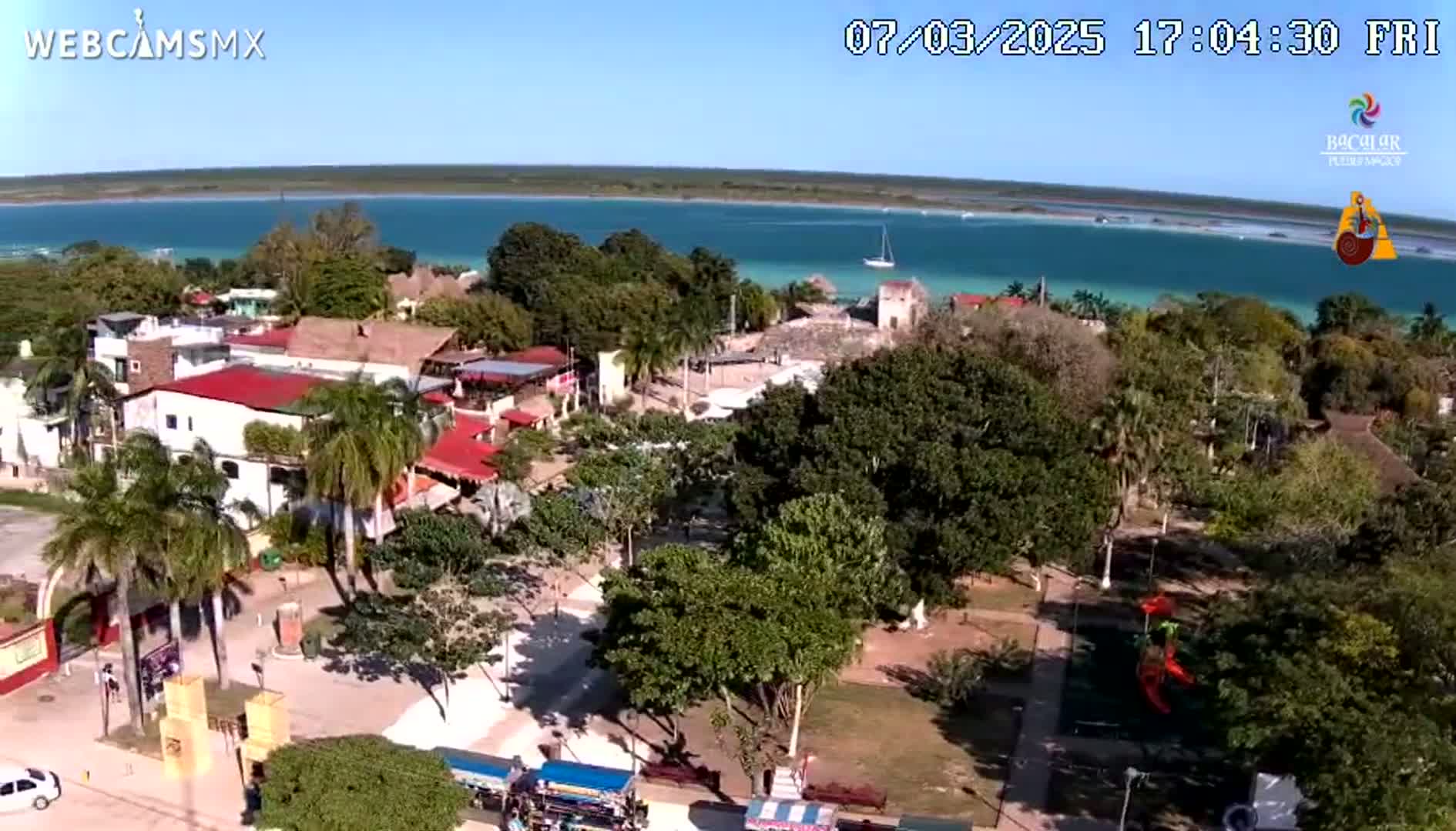An aerial view shows a town with lush green trees and buildings near a calm, turquoise body of water under a clear, sunny sky.