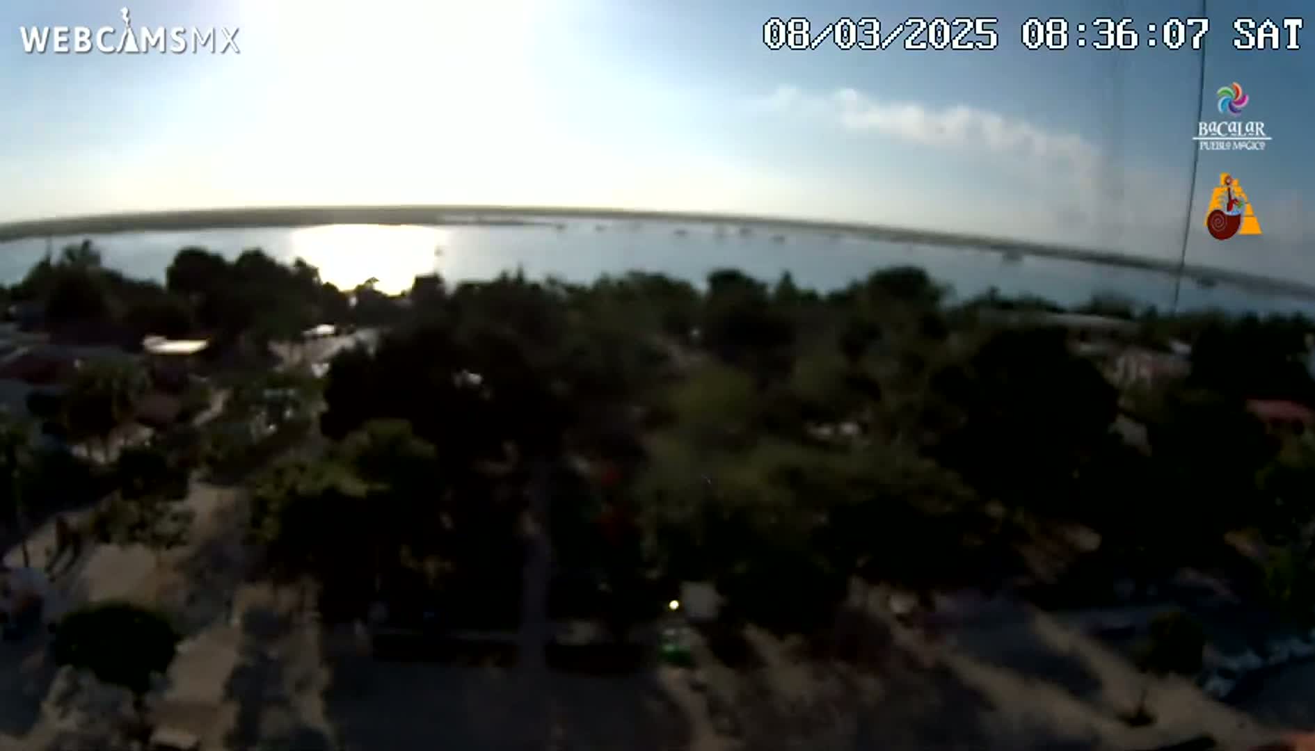 An elevated view shows a calm body of water reflecting the sunrise, with a town and lush green vegetation in the foreground under a mostly clear sky.