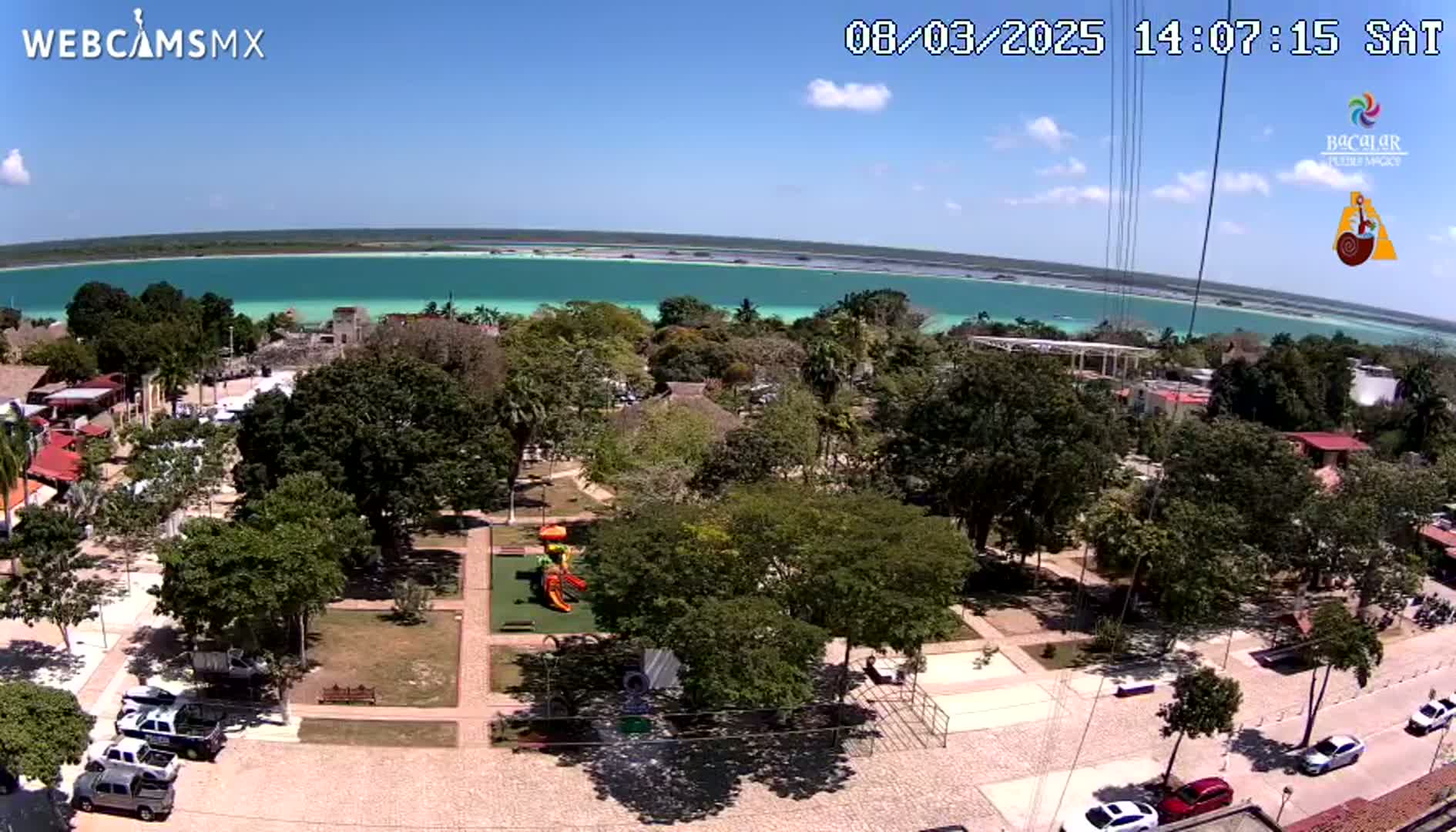 An aerial view shows a park with a playground next to a town bordering a bright turquoise body of water under a sunny sky.