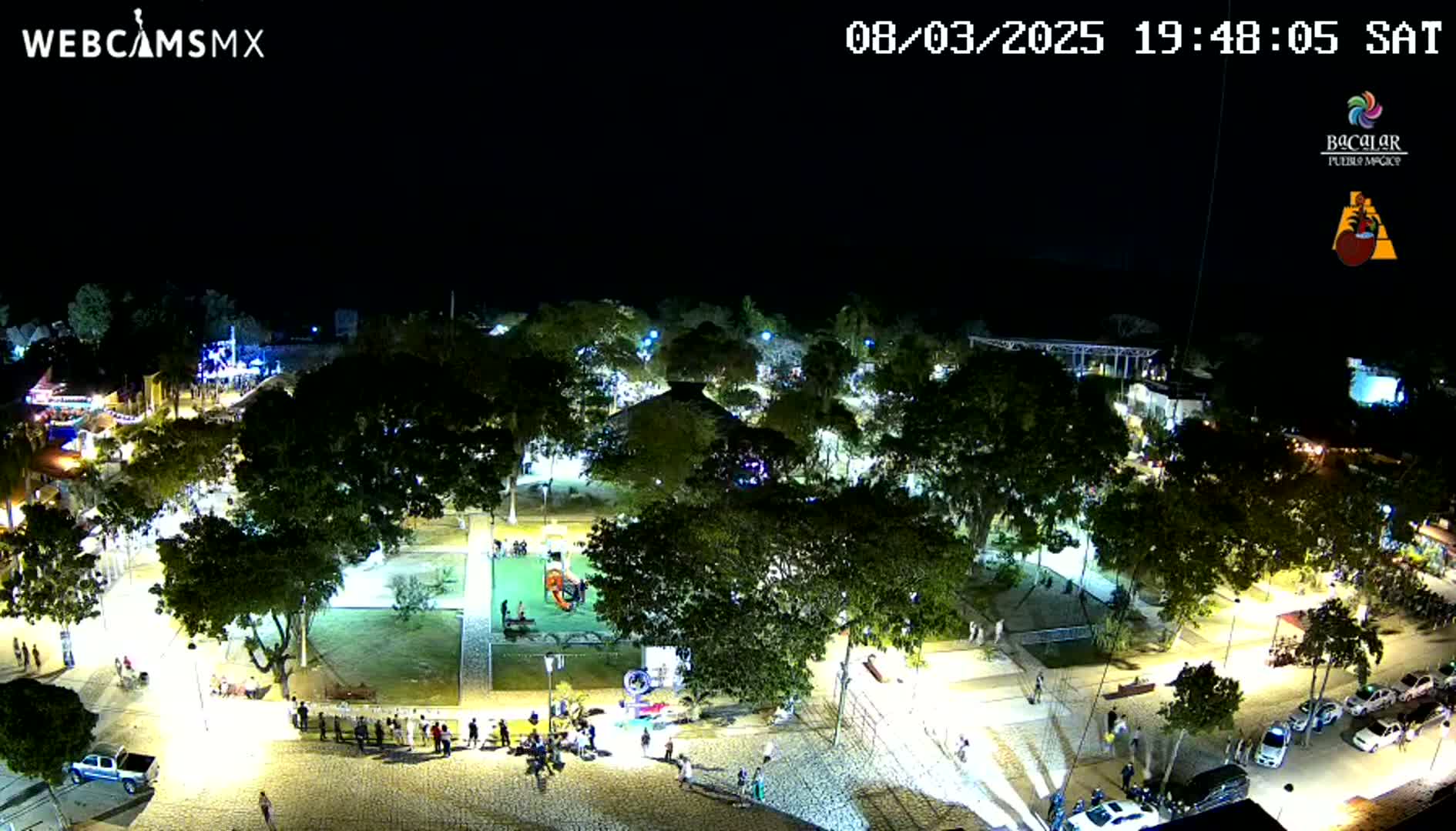 A nighttime, aerial view of a town square with many trees, people, and cars, illuminated by lights.