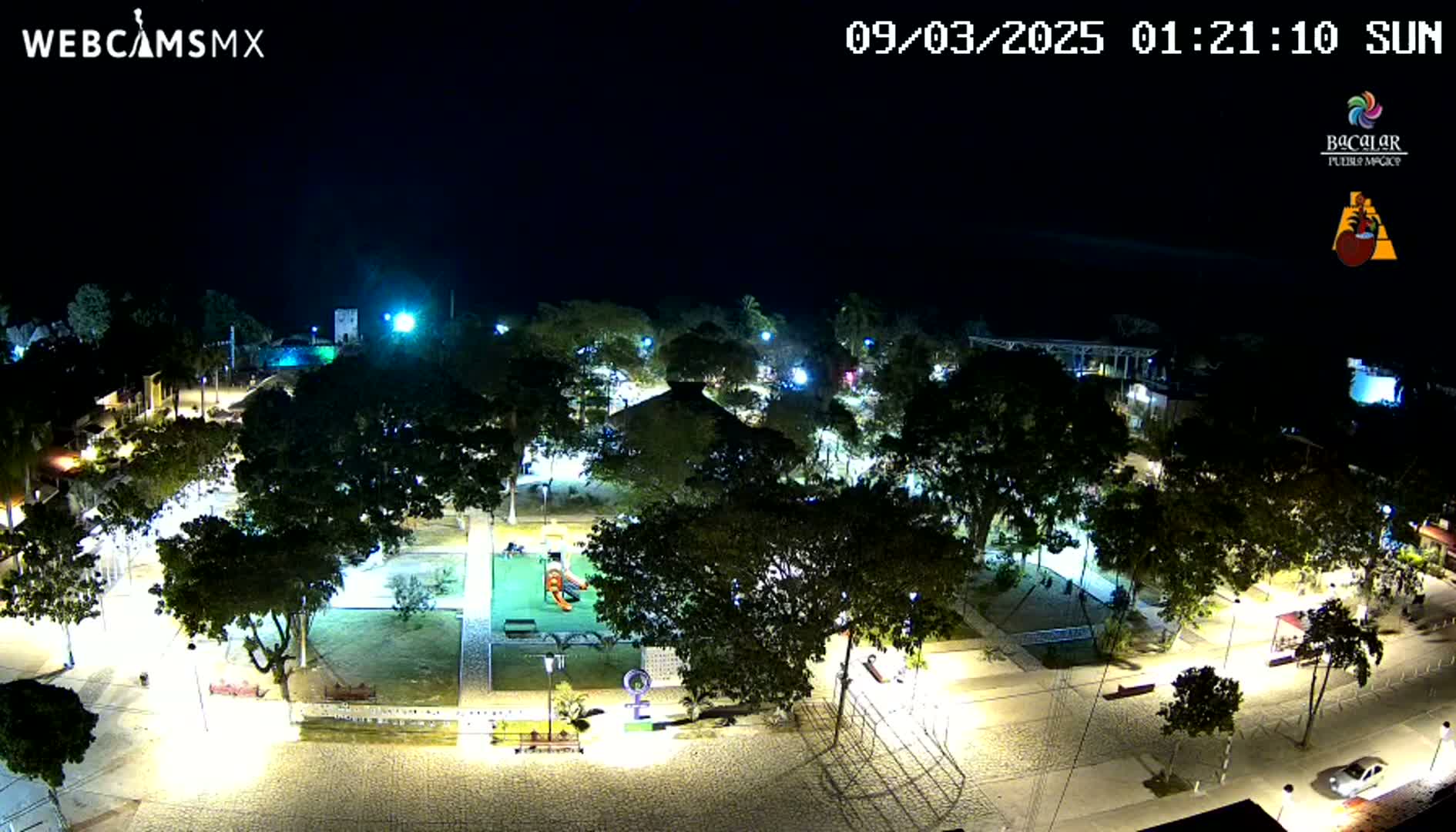 A brightly lit town square at night, filled with lush trees and a playground, under a clear, dark sky.