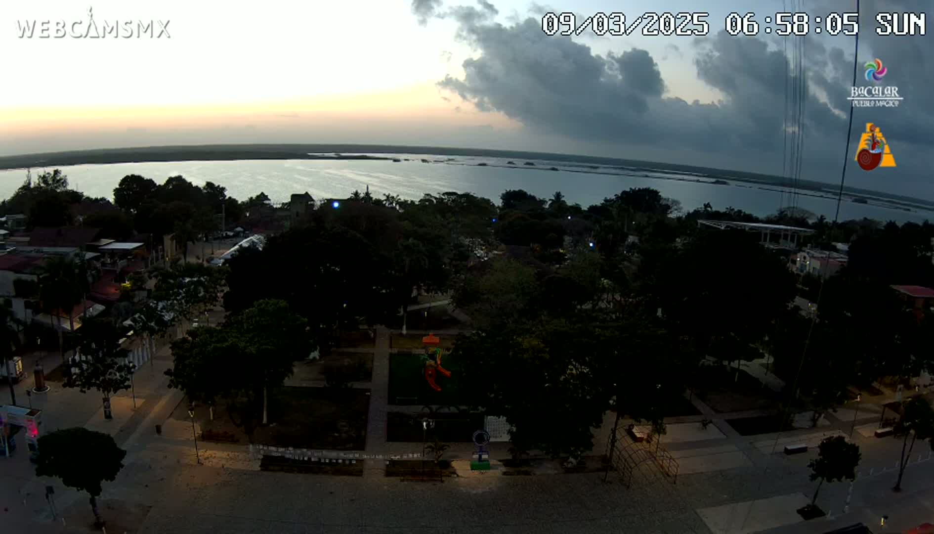 An aerial view at dusk shows a town square with a playground and surrounding buildings, next to a calm body of water under a partly cloudy sky.