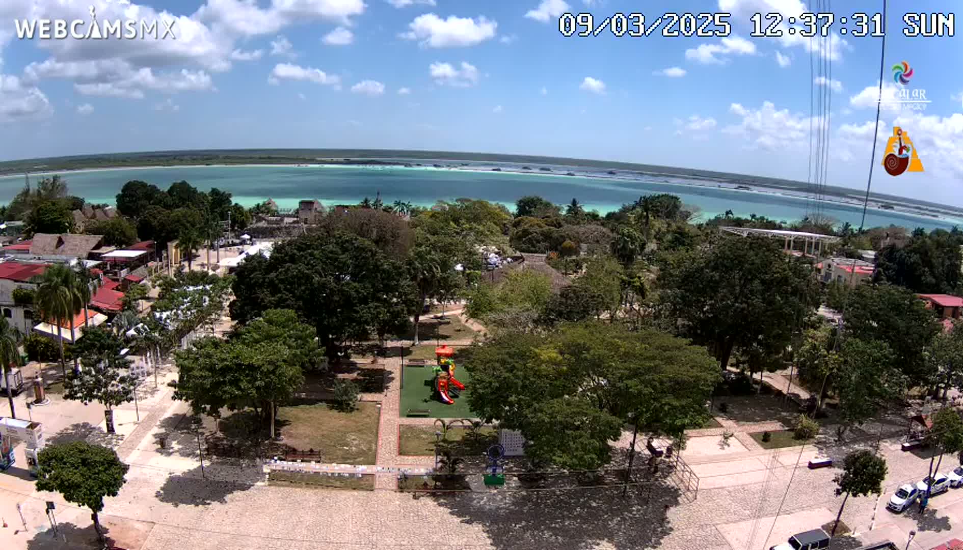 A sunny day overlooking a town square with a playground, surrounded by lush green trees and a calm, turquoise body of water in the background.