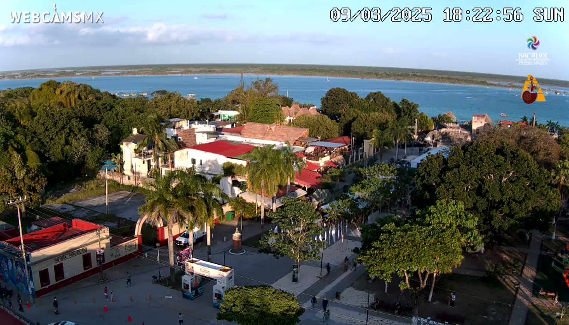 An aerial view shows a town next to a calm, blue body of water under a mostly clear, sunny sky.