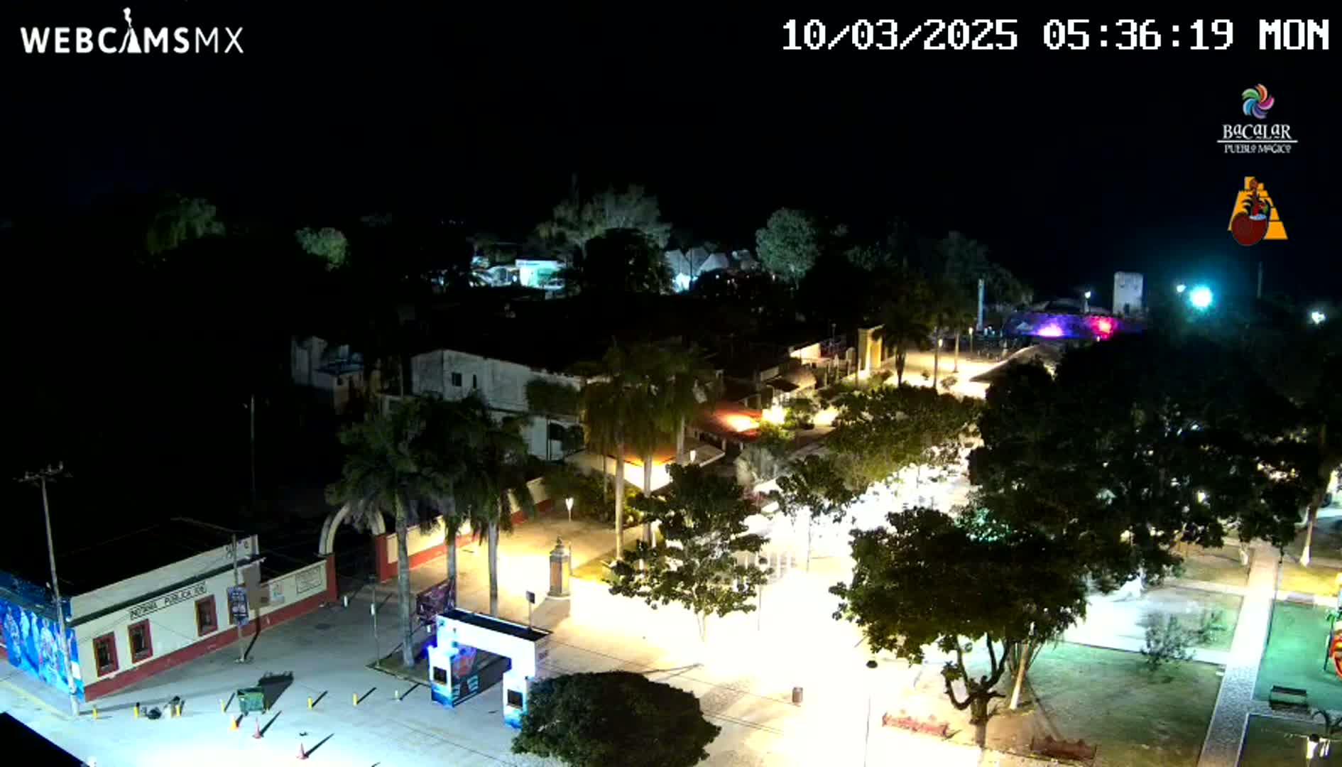 A nighttime aerial view of a town square, illuminated by streetlights, with palm trees and buildings surrounding it under a clear sky.