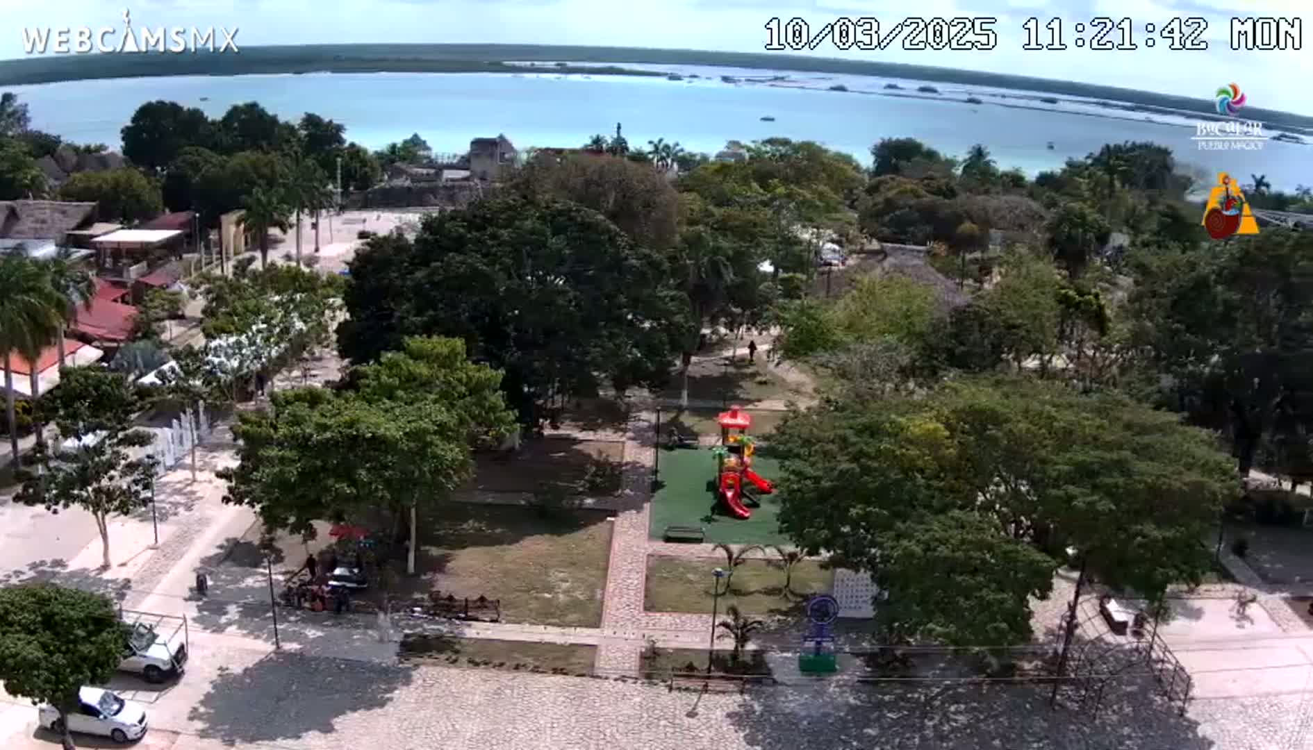 An aerial view shows a town square with lush green trees, a playground, and a body of calm, blue water visible in the background under a sunny sky.